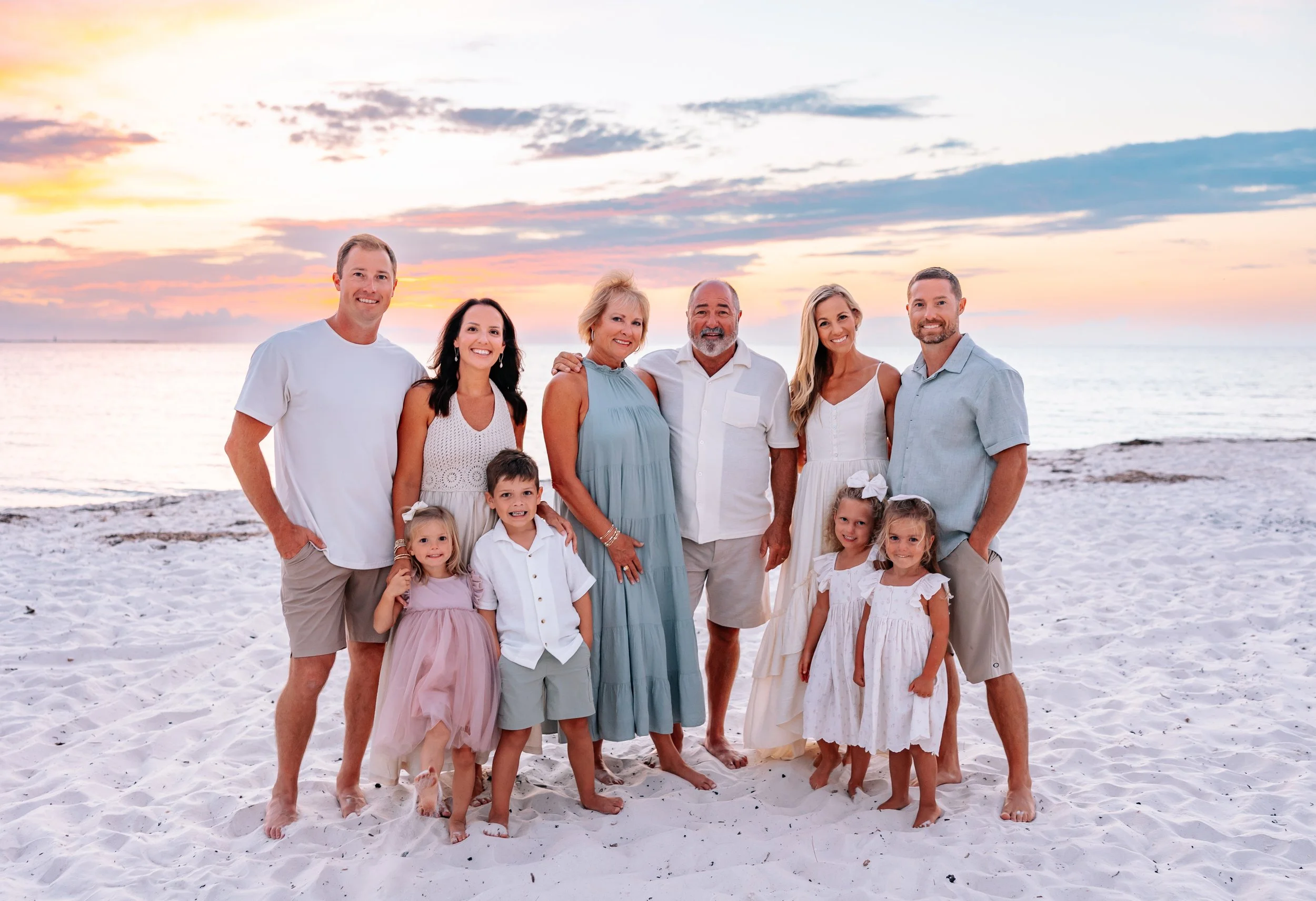 Family group standing on a beach at sunset, smiling, with the ocean and colorful sky in the background.