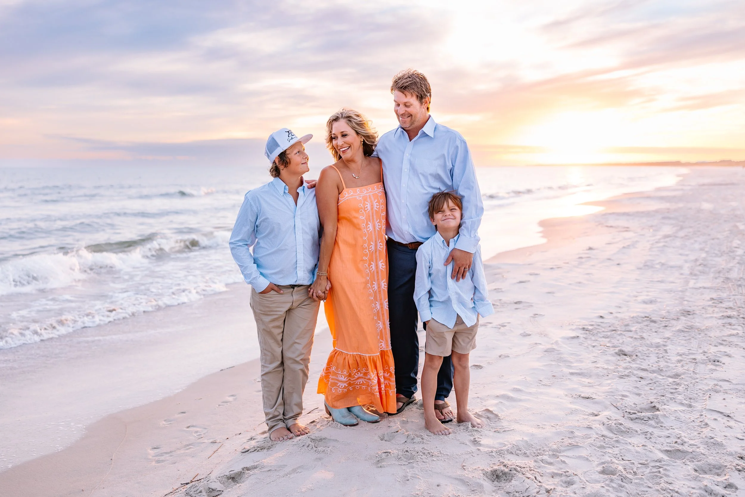 A family of four standing on the beach at sunset, smiling and looking at each other. The ocean waves are behind them, and they are dressed in casual summer clothes.