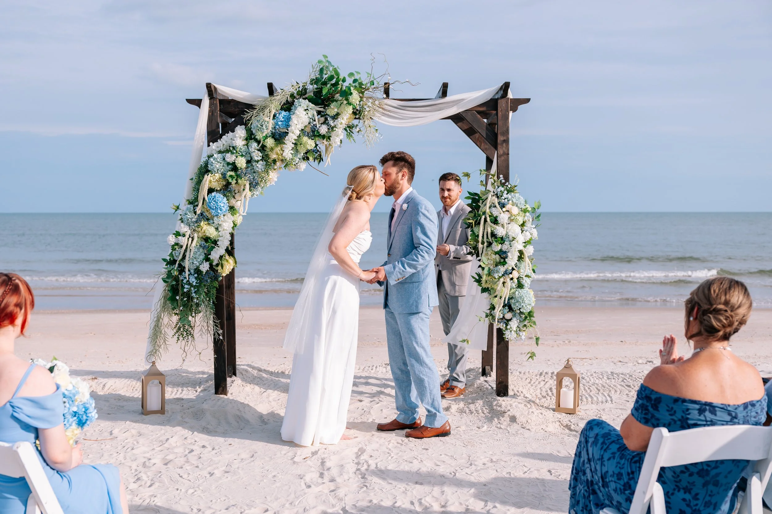 A couple kissing during a beach wedding ceremony under a wooden arch decorated with white, blue and green flowers and draped fabric, with guests seated on white chairs along the sandy shore, ocean and sky in the background.