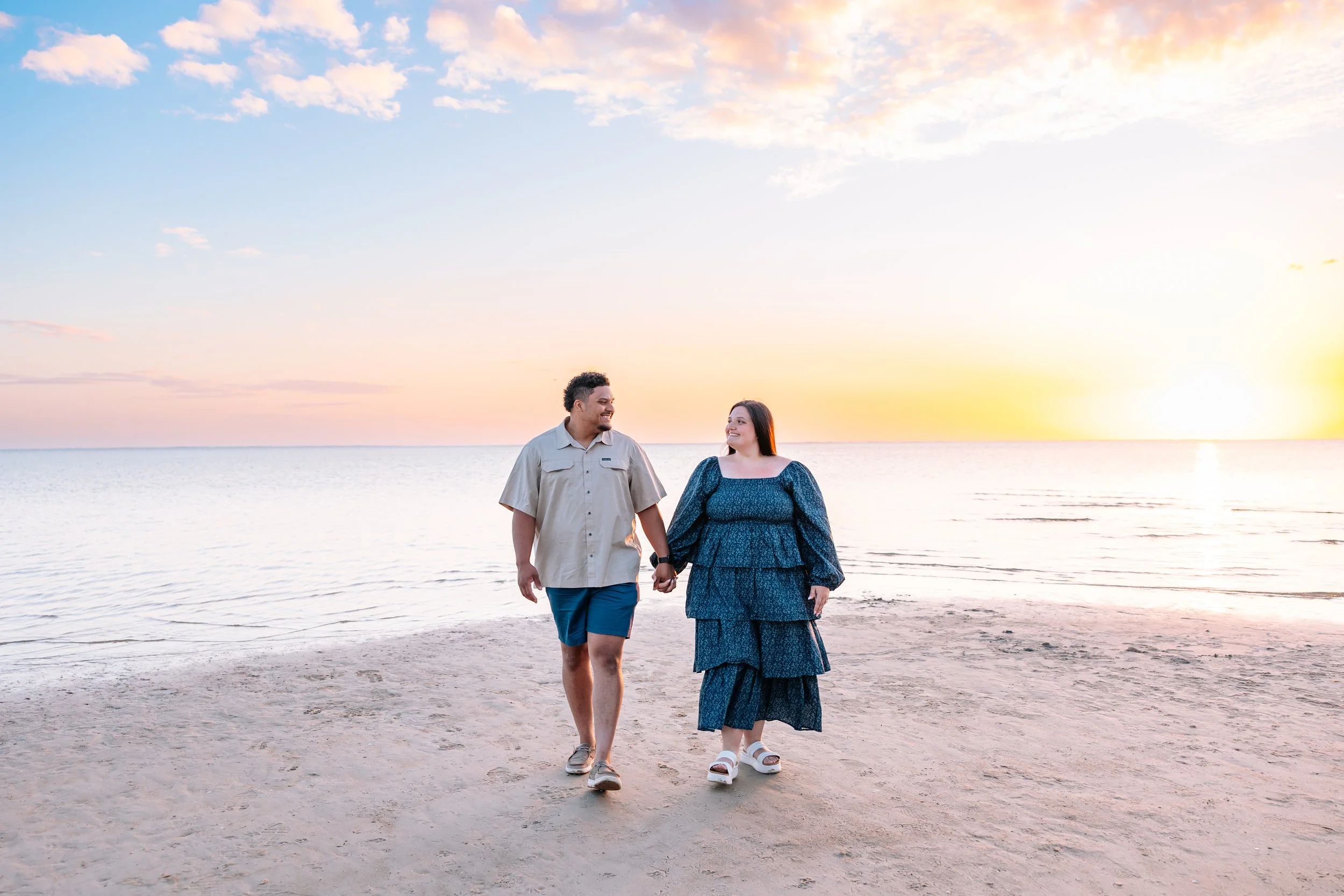 A couple walking hand in hand on a beach during sunset, smiling and looking at each other, with gentle waves in the background and a partly cloudy sky.