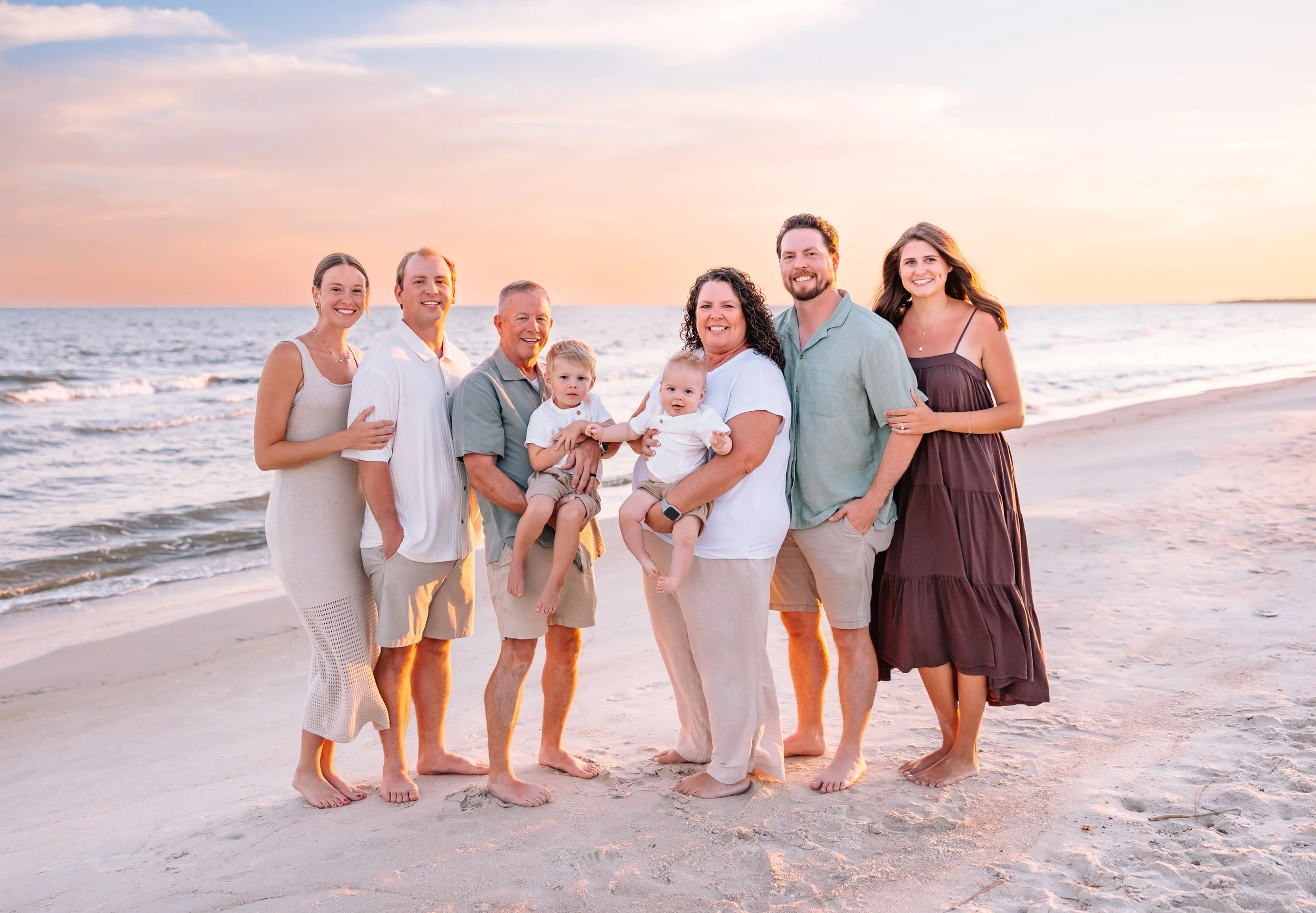 A family of nine people standing on the beach at sunset, smiling, with the ocean in the background.