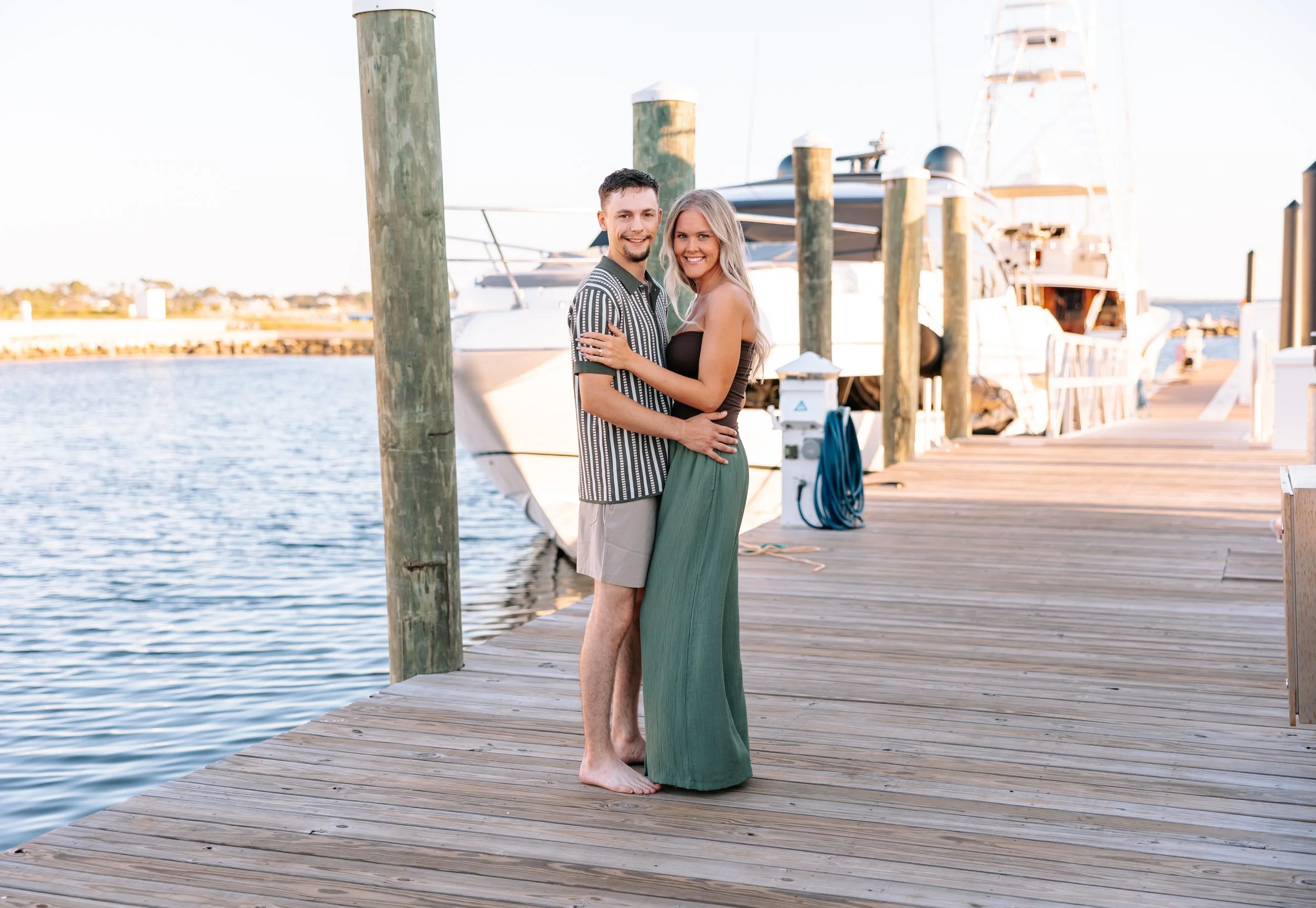 A couple standing on a wooden dock by the water, smiling at the camera, with a yacht in the background.
