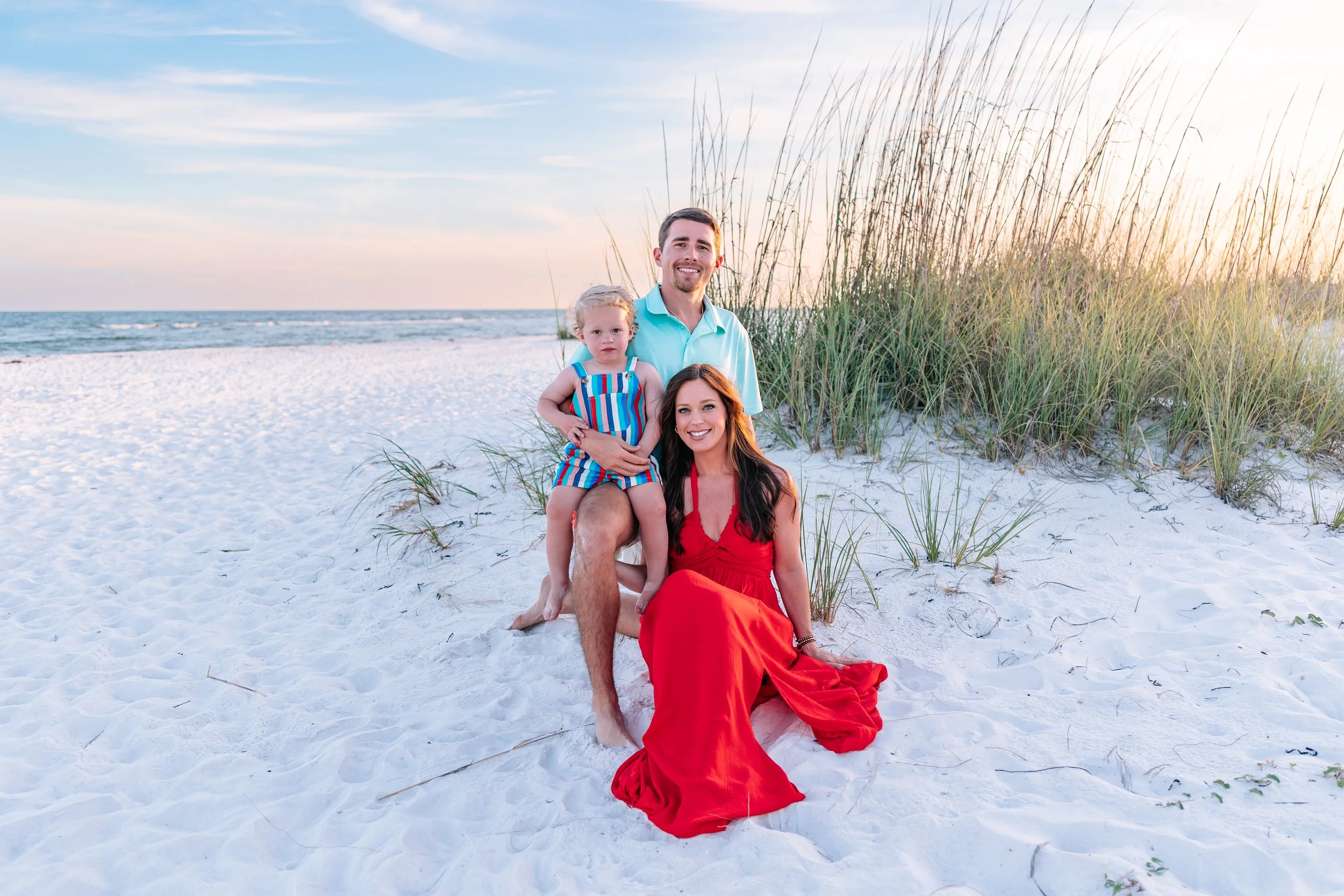A family of three, a man, a woman, and a young girl, sitting on white sandy beach with tall grass and ocean in the background during sunset. The woman is in a red dress, the man is in a light blue shirt, and the girl wears a colorful striped outfit.