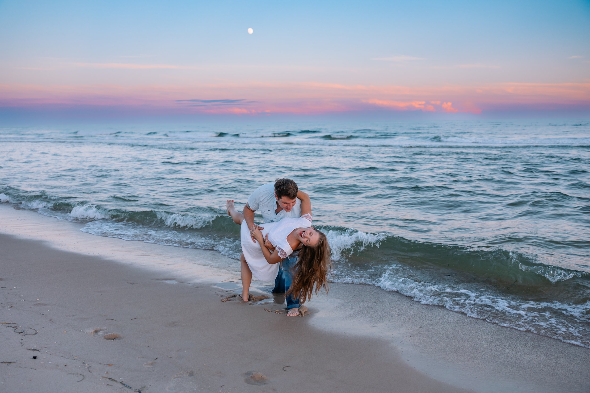 A young man and woman are at the beach, the man is holding the woman upside down by her waist as she laughs. They are near the shoreline with waves washing up onto their feet. The sky has pink and blue hues with the moon visible.