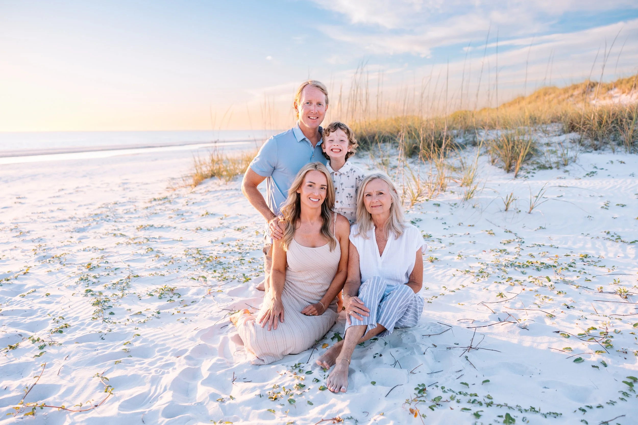 A multi-generational family of five sitting and standing on a sandy beach with dune grass, during sunset or sunrise, smiling and enjoying their time together.