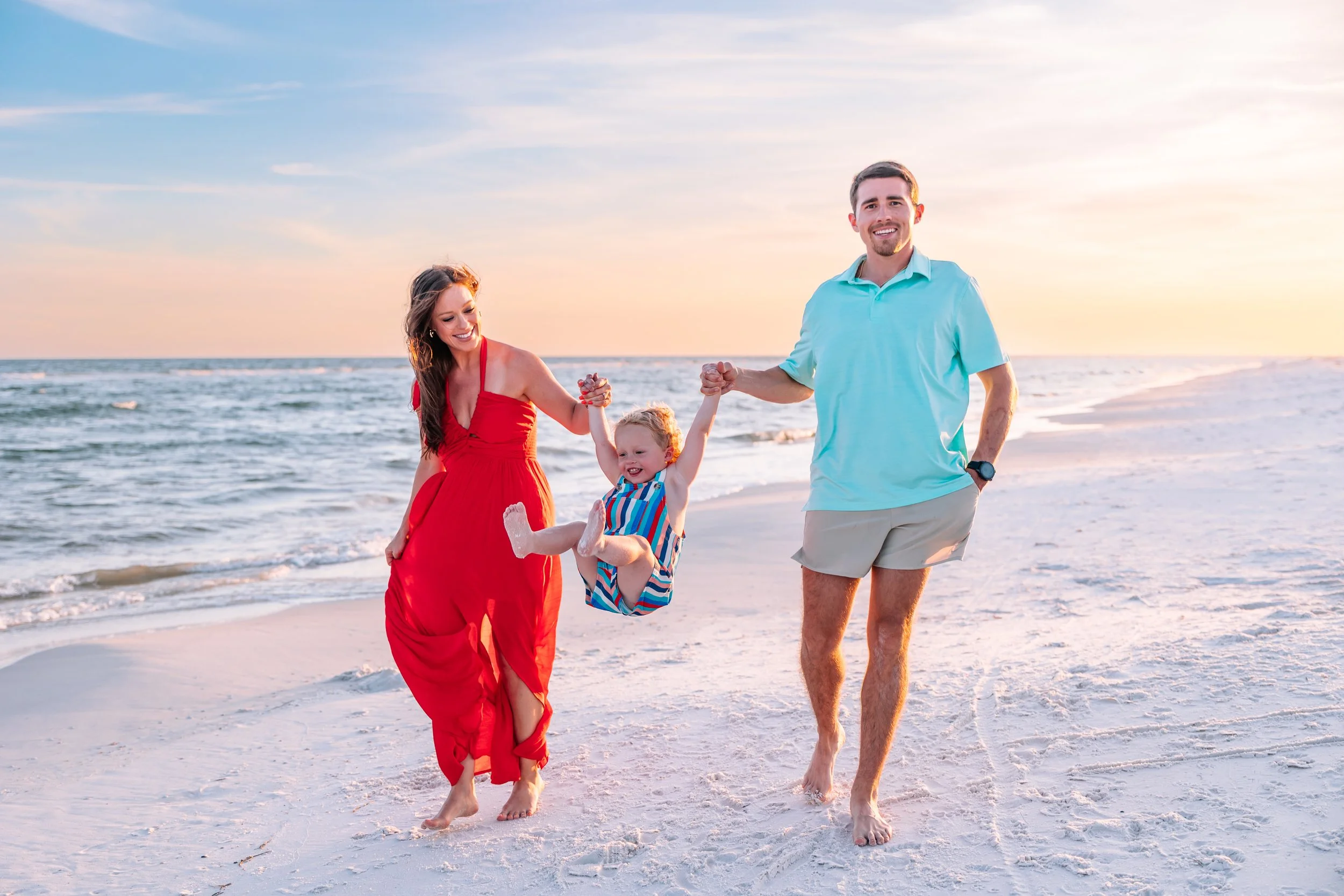 A family of three with a woman in a red dress, a man in a light blue shirt, and a young child in striped clothing, walking barefoot on the beach at sunset, holding hands and smiling.