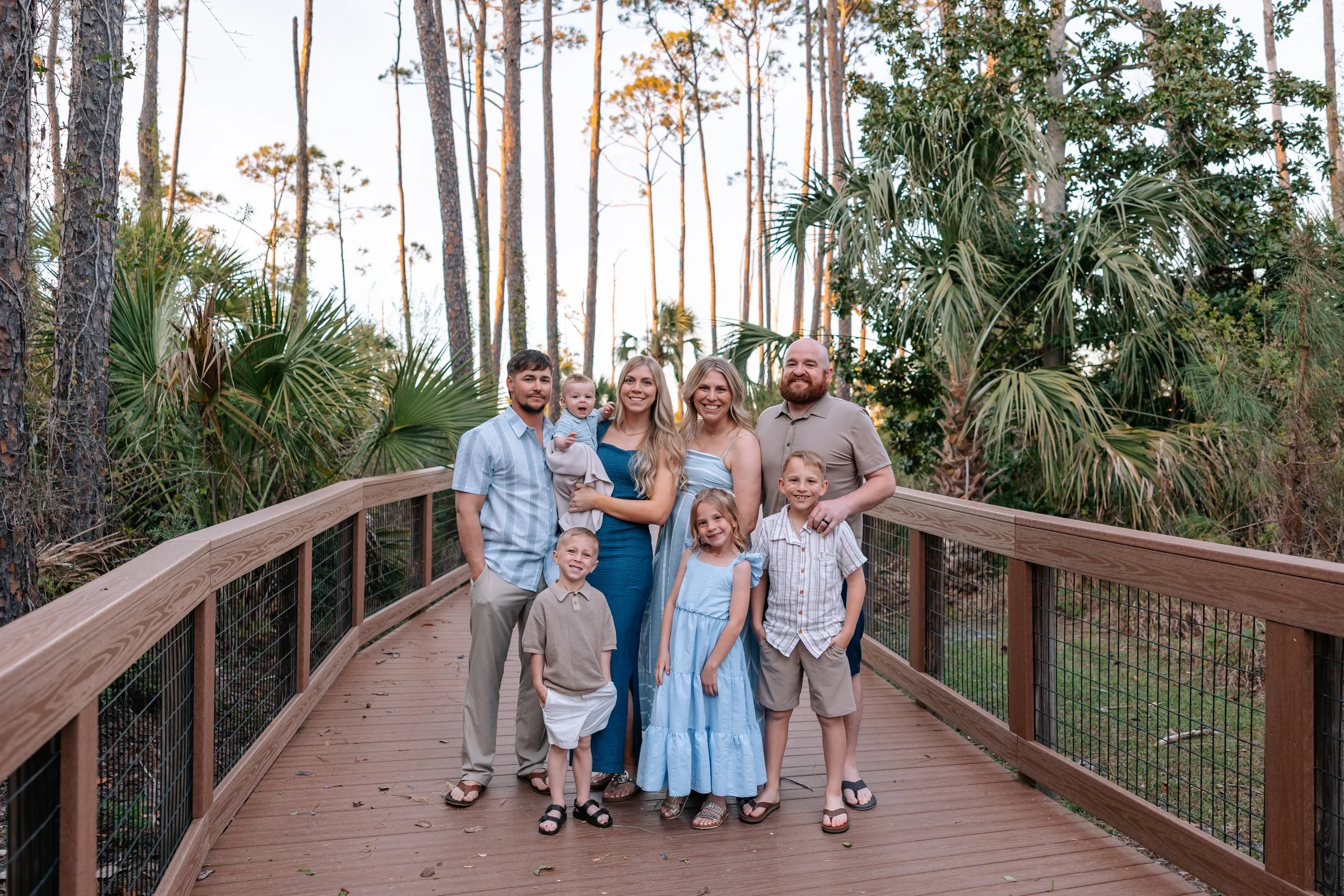 A group of nine people, including children, standing on a wooden bridge in a forest with tall trees and lush greenery, smiling and posing for a photo.