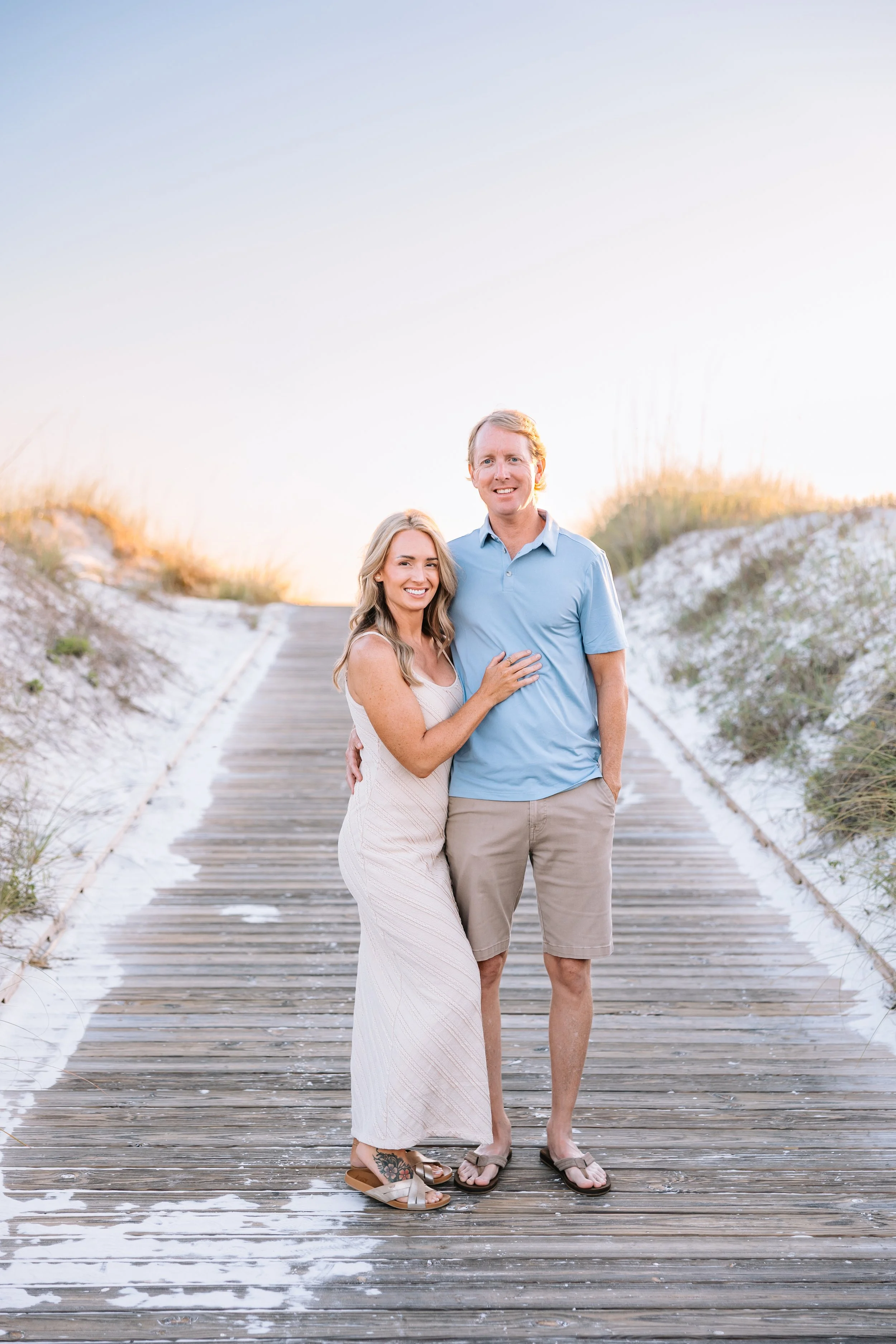 A smiling couple stands close together on a wooden boardwalk, with the woman wearing a white dress and the man wearing a light blue shirt and khaki shorts, with a sandy beach and grassy dunes in the background during sunset.
