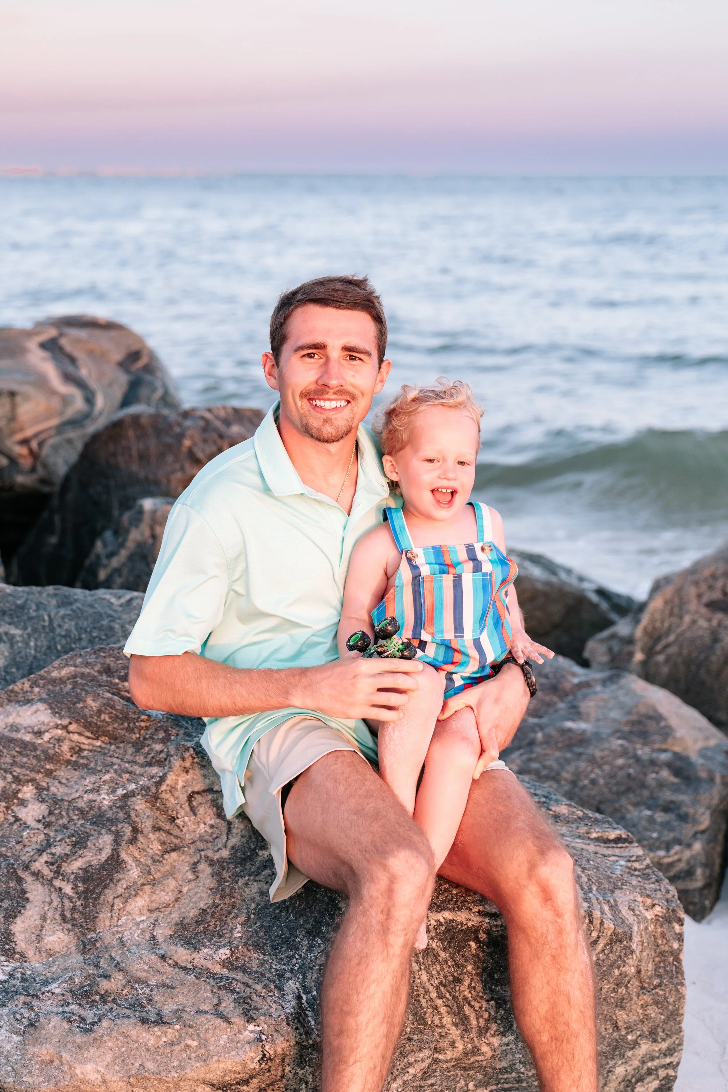A man with short brown hair and a beard, wearing a light green polo shirt, sitting on rocks at the beach, holding a young boy with blonde curly hair wearing a colorful striped tank top, both smiling with the ocean and sky in the background.