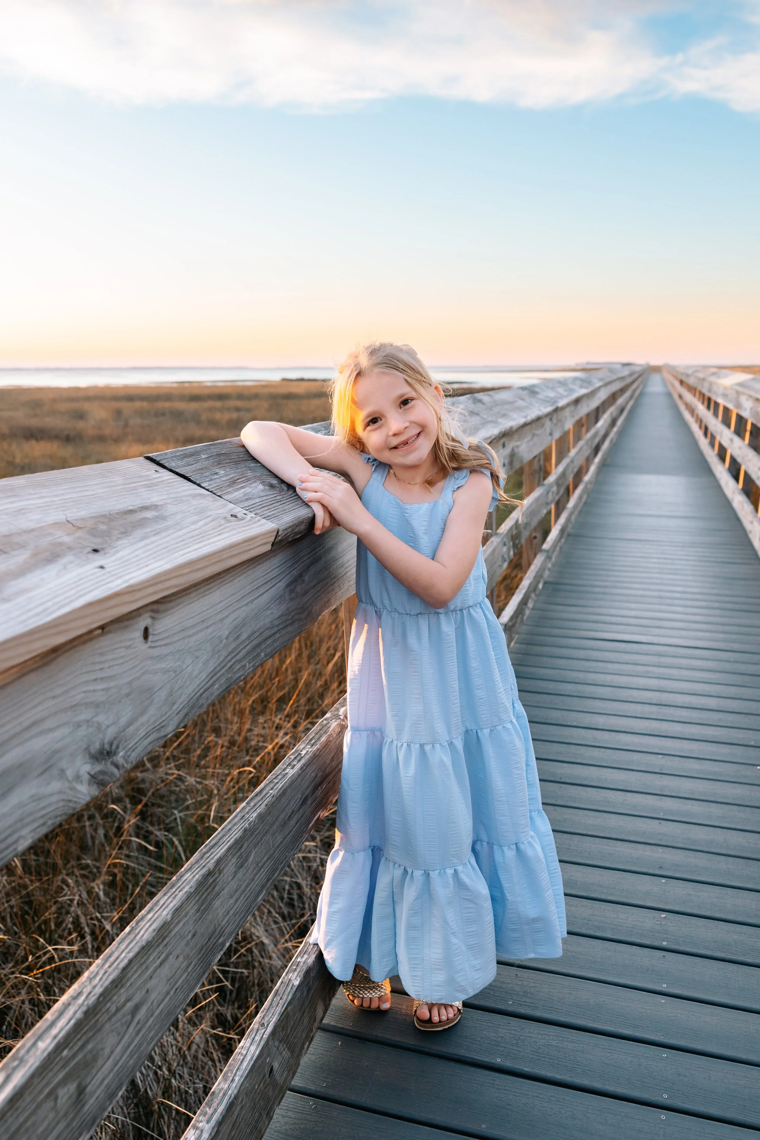 A young girl in a light blue sleeveless dress smiling and leaning on a wooden railing on a boardwalk during sunset, with an open landscape and water in the background.