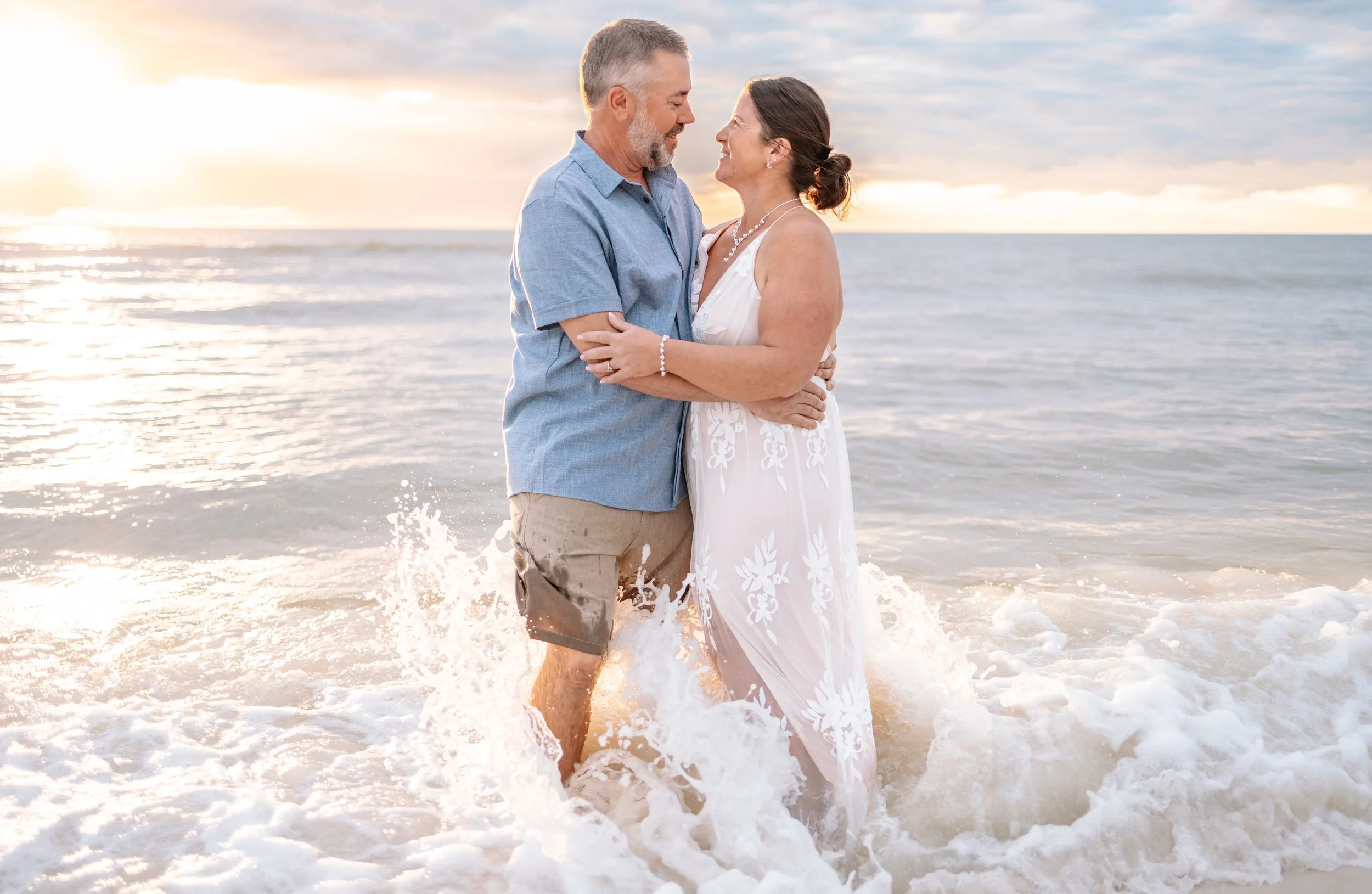 A couple standing close and smiling at each other on a beach during sunset, with the ocean waves gently crashing around their legs.