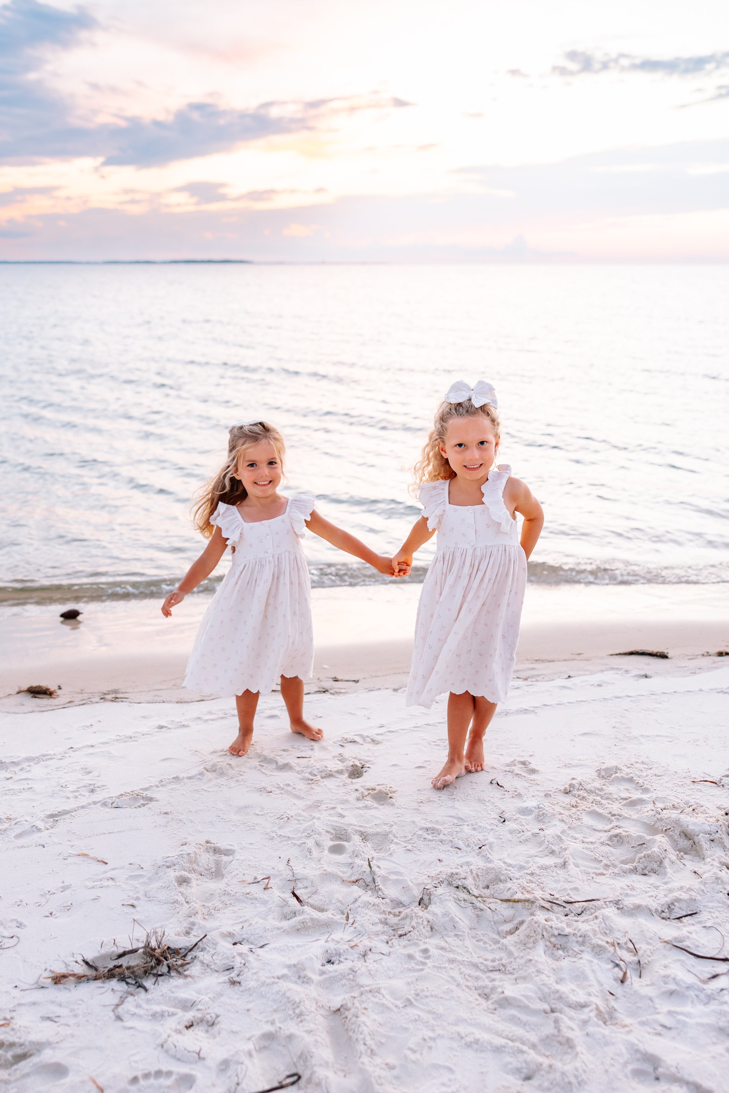 Two young girls in white dresses holding hands and walking on a sandy beach by the ocean during sunset
