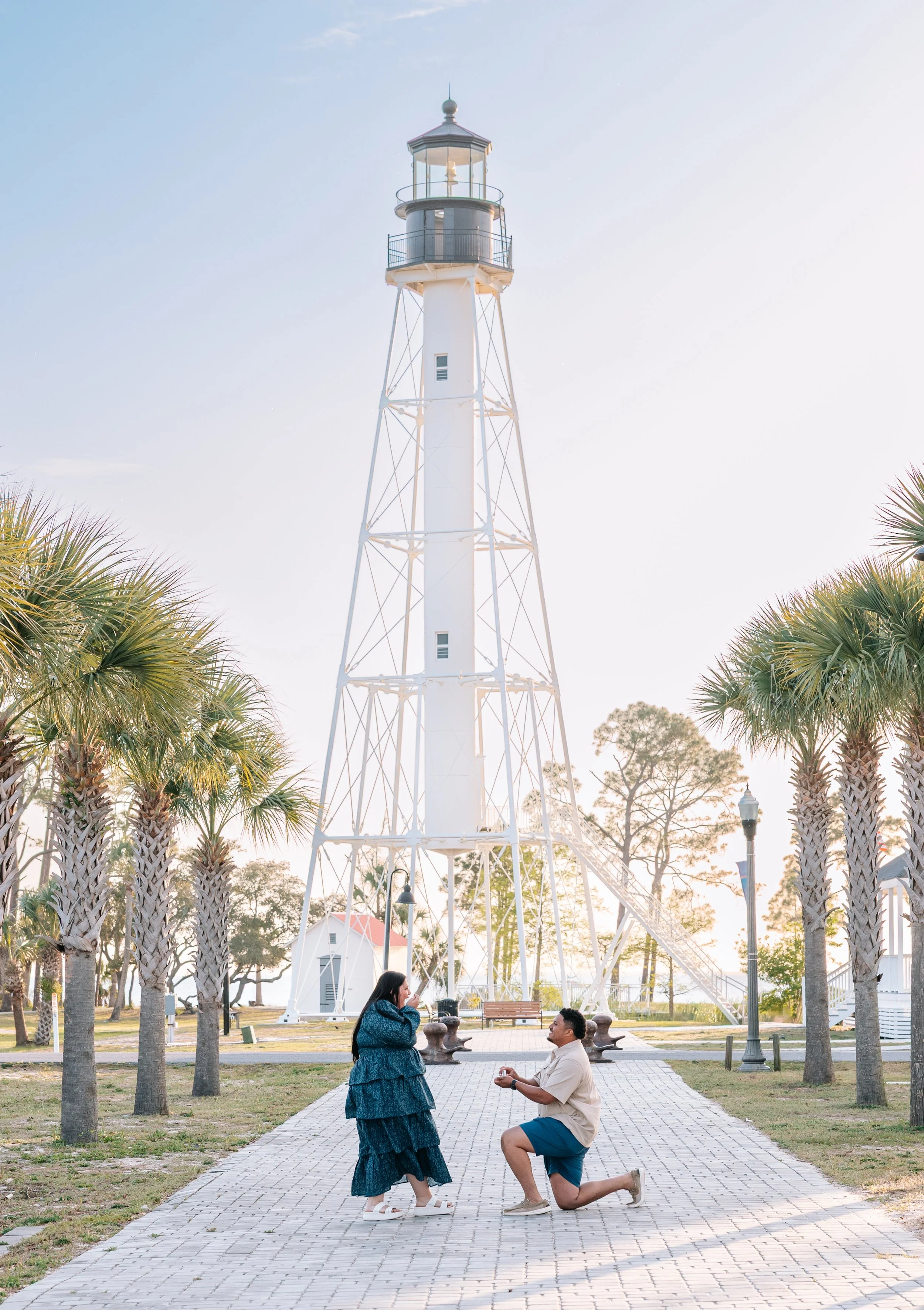 A couple dressed in semi-casual clothing, with the man on one knee proposing to the woman in a park with palm trees and a white lighthouse in the background during sunset.