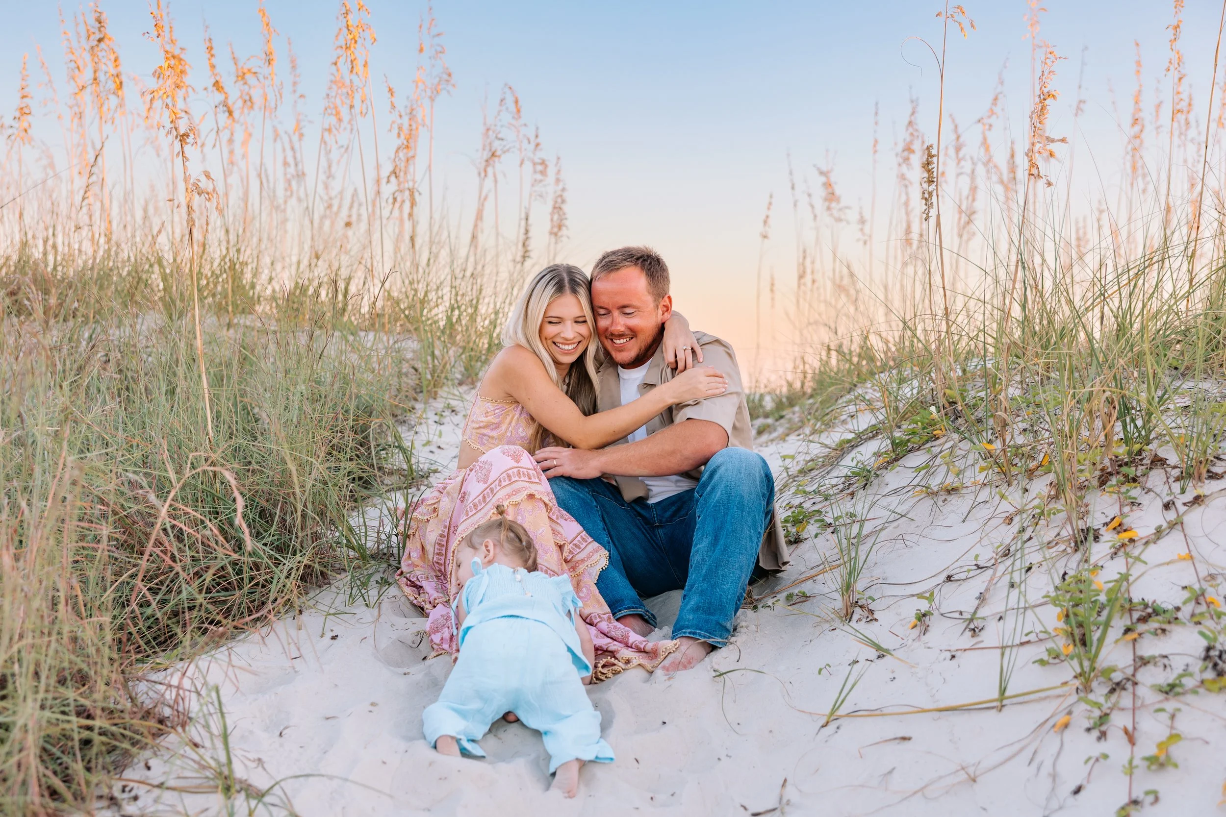 A couple sitting on the sandy beach, smiling and hugging each other, with a small girl laying on the sand in front of them, surrounded by tall beach grass and a clear sky with a sunset.