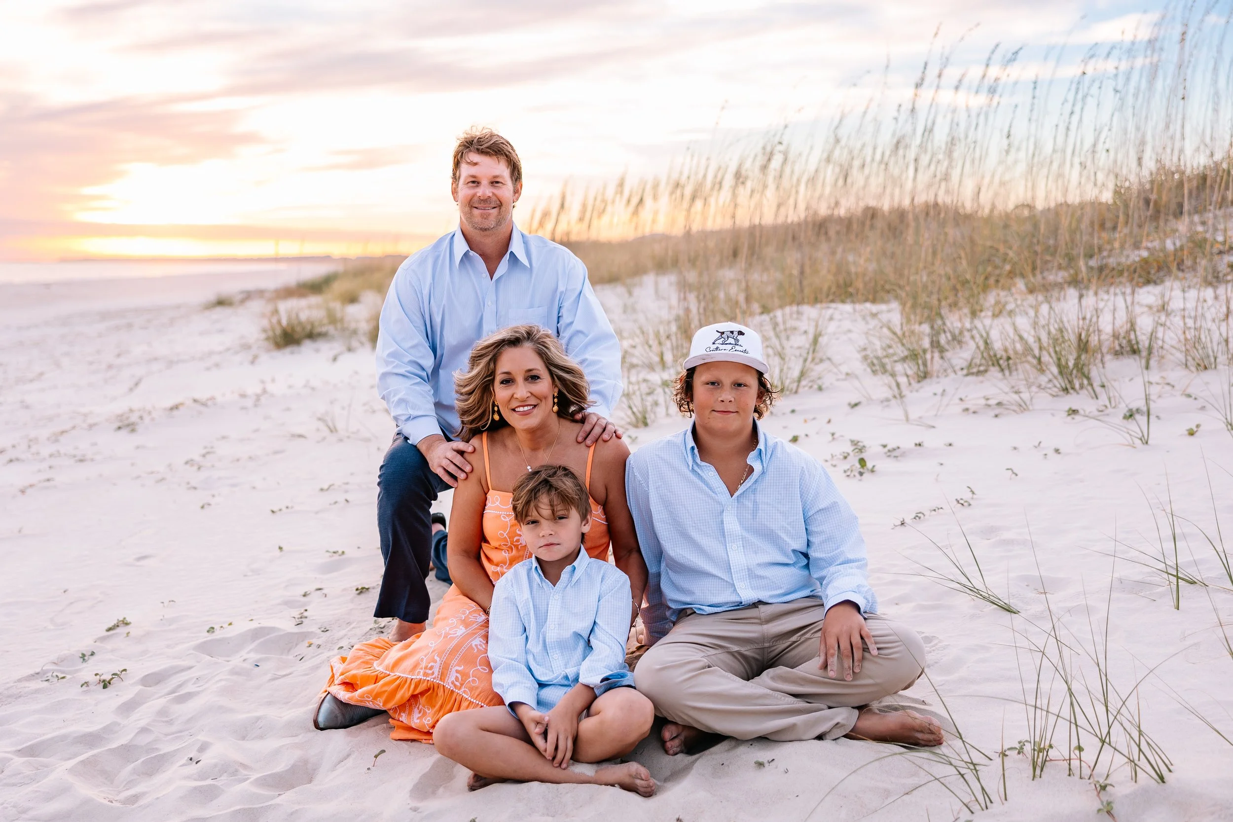 A family of five posing on a sandy beach at sunset. The family includes a man, a woman, two boys, and a girl, all dressed in casual summer clothes. They are sitting and kneeling on the sand with dunes and beach grass in the background.