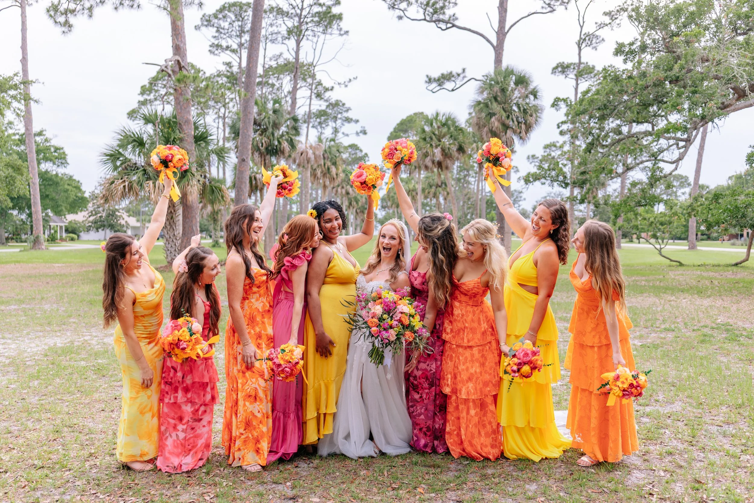 A group of women in colorful dresses holding bouquets of flowers, celebrating outdoors in a park with trees.