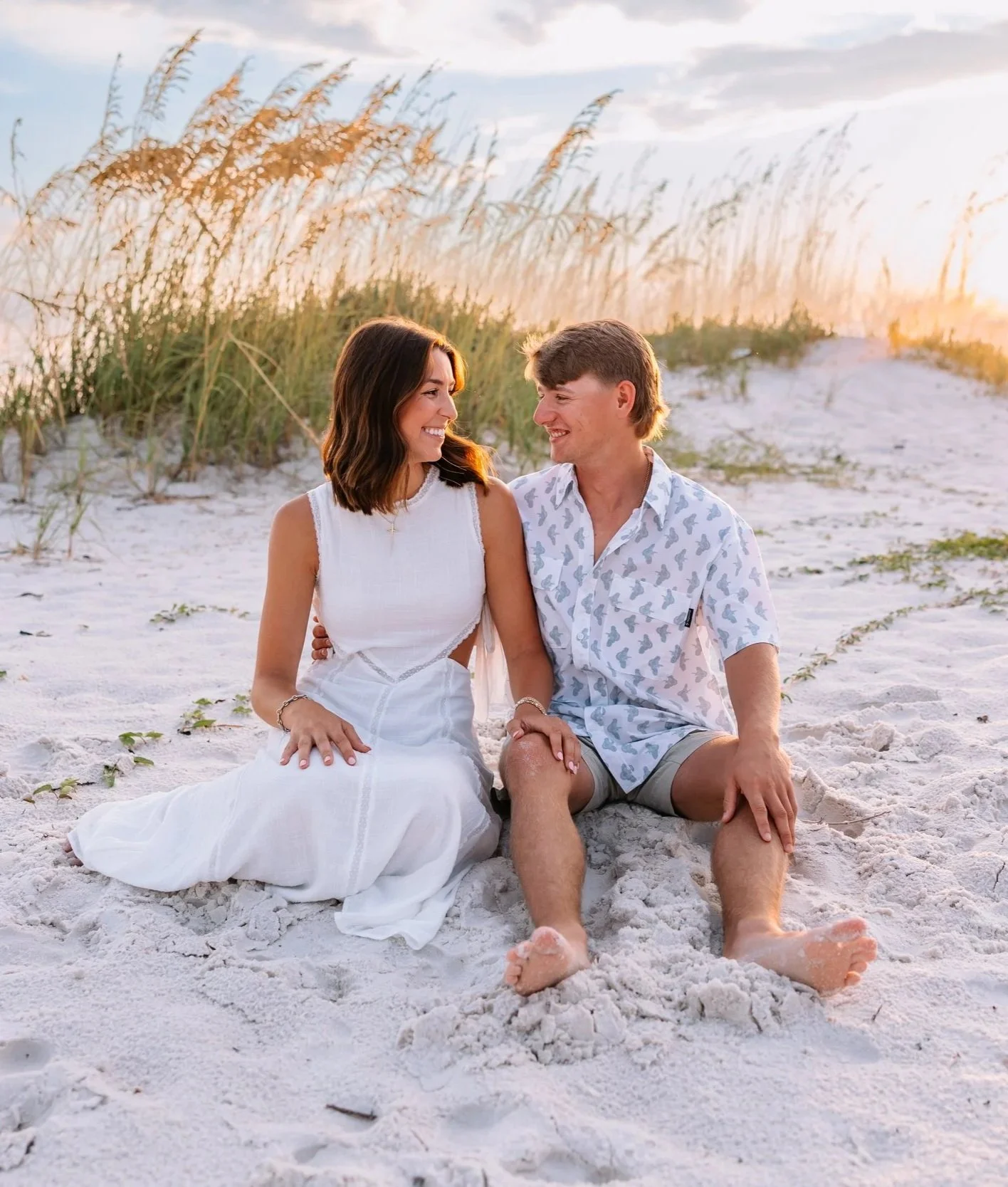 A smiling woman and man sitting on a sandy beach at sunset, looking at each other.