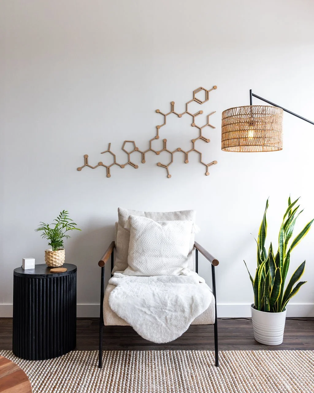 Modern living room with a beige armchair, black side table with a plant, large potted snake plant, minimalist wall art resembling chemical structures, woven pendant lamp, and a beige and white rug.