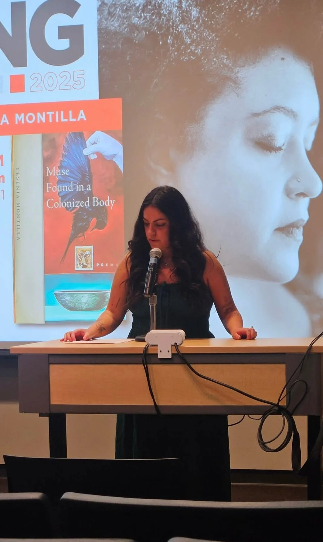 A woman with long dark hair stands at a podium, speaking into a microphone during a presentation or reading event. Behind her, a large projected image features a close-up profile of a woman's face and a book cover titled "Muse Found in a Colonized Body." The setting classroom.