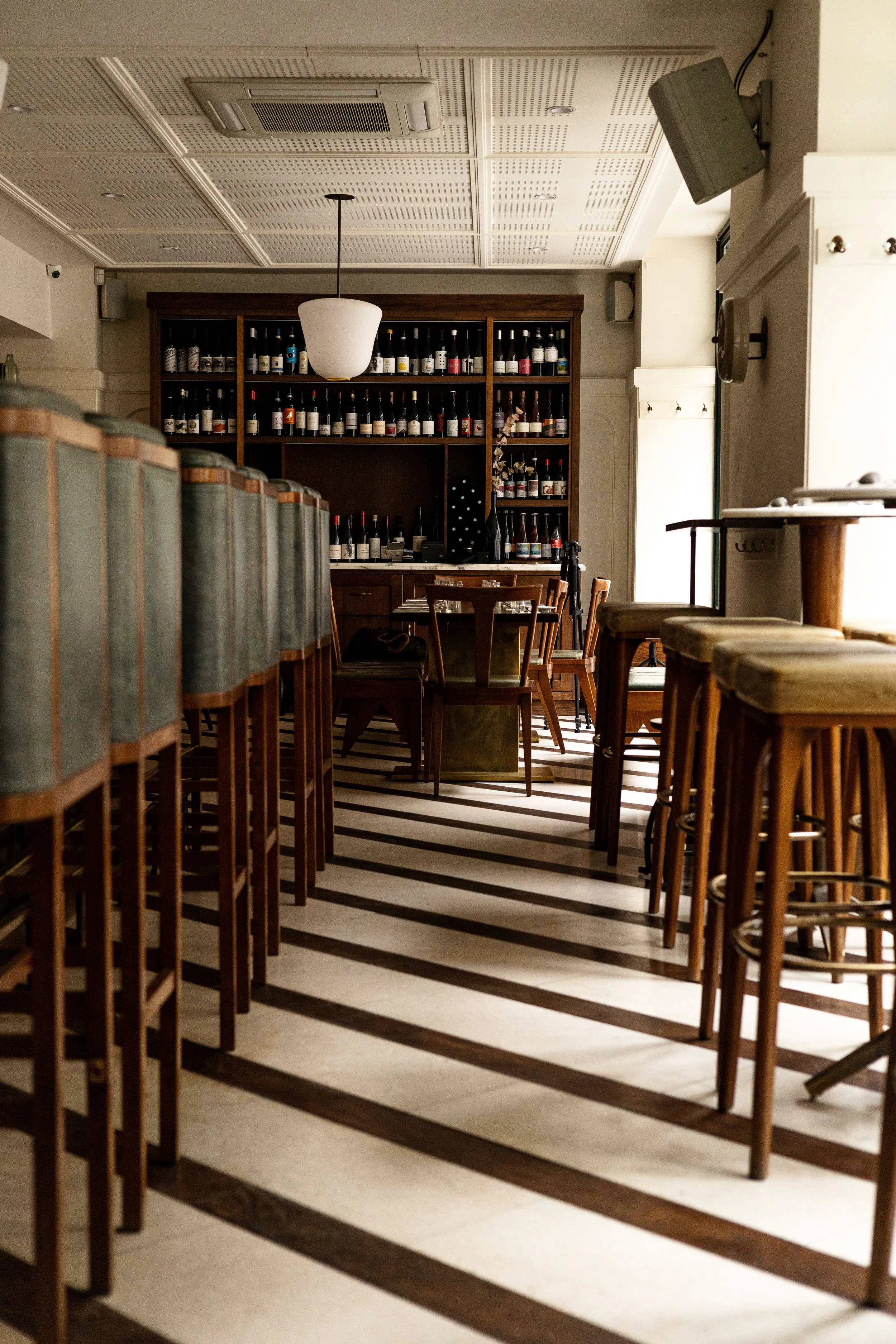 Intérieur d'un restaurant ou bar avec chaises hautes en bois, étagère avec bouteilles de vin, tables et chaises, plafond en panneaux, lumière naturelle venant d'une fenêtre.