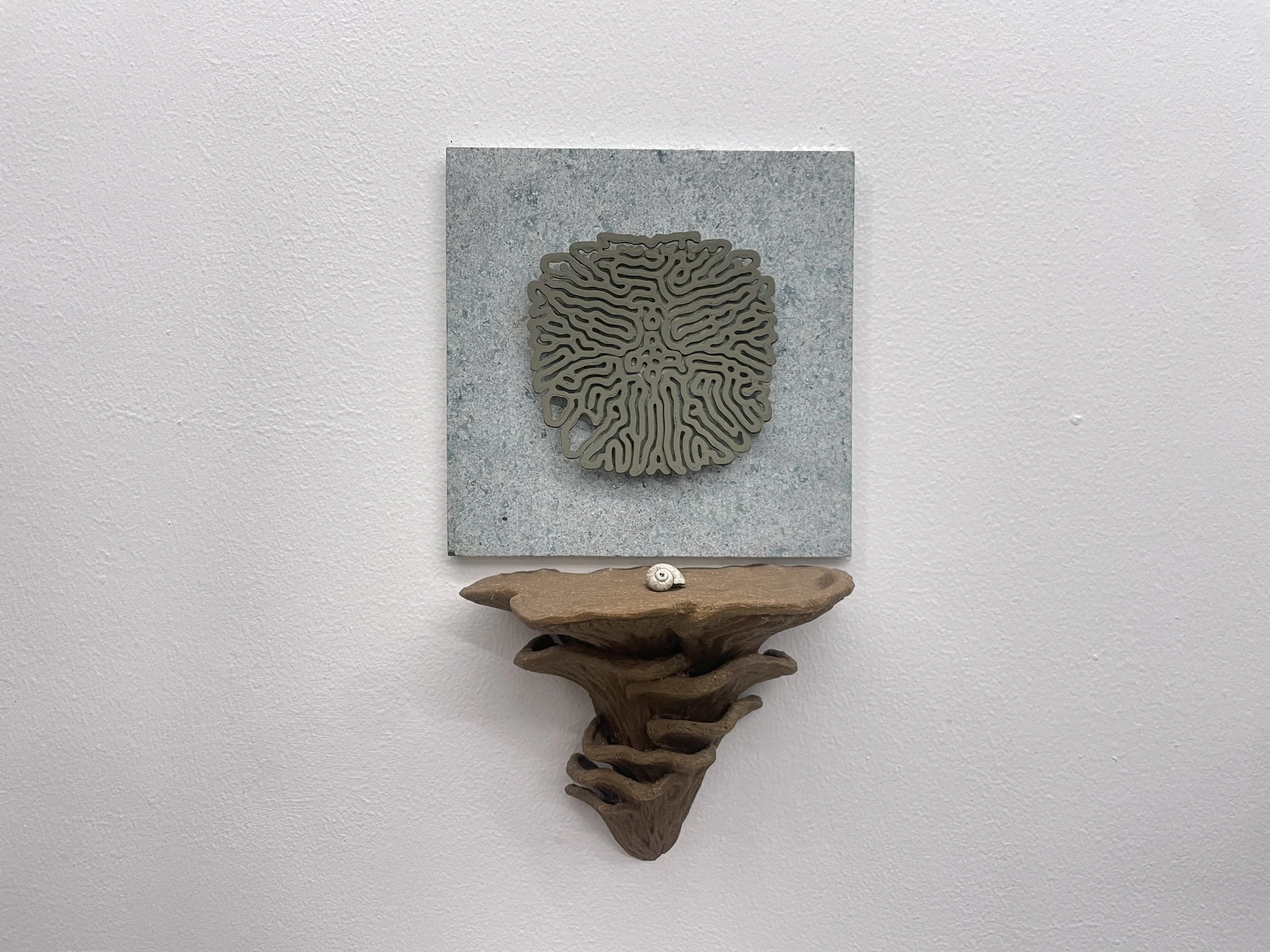 Mixed-media art piece mounted on a white wall, featuring a metal maze-like sculpture, a square stone or concrete background, and a wooden structure resembling a sea shell or coral. Hypha Inc Marketing Agency and Creative Studio Lab, Tulum.