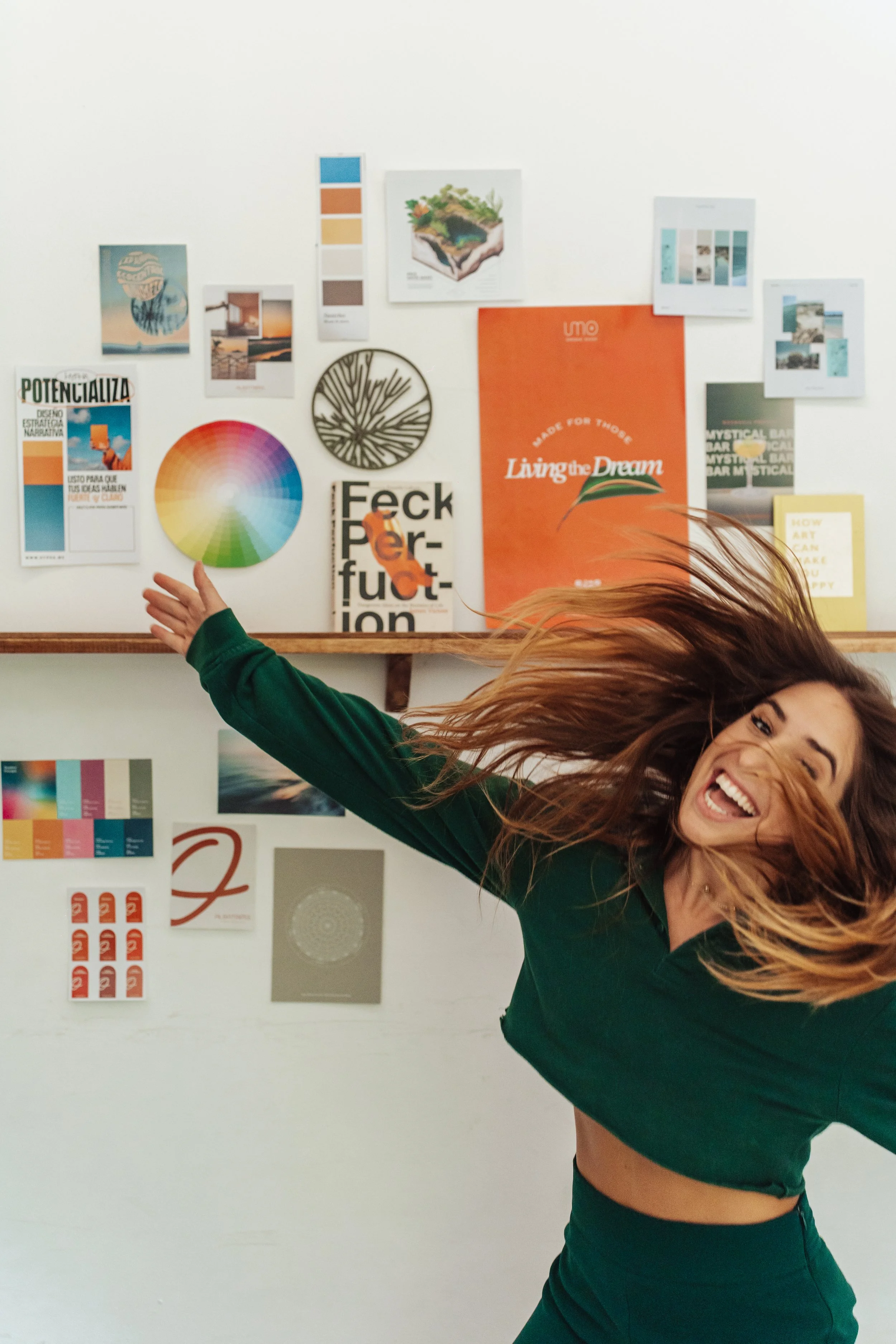 A woman wearing a green outfit, smiling and jumping with her hair flying, in front of a white wall decorated with various colorful posters and prints. APOD Hypha