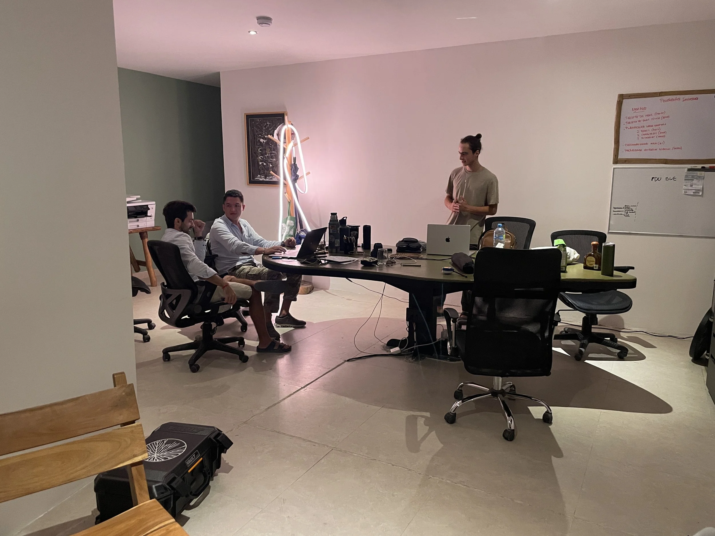 Three men sitting at a large conference table with laptops, and another man standing near a whiteboard in a modern office space. Hypha Inc Marketing Agency and Creative Studio Lab, Tulum.