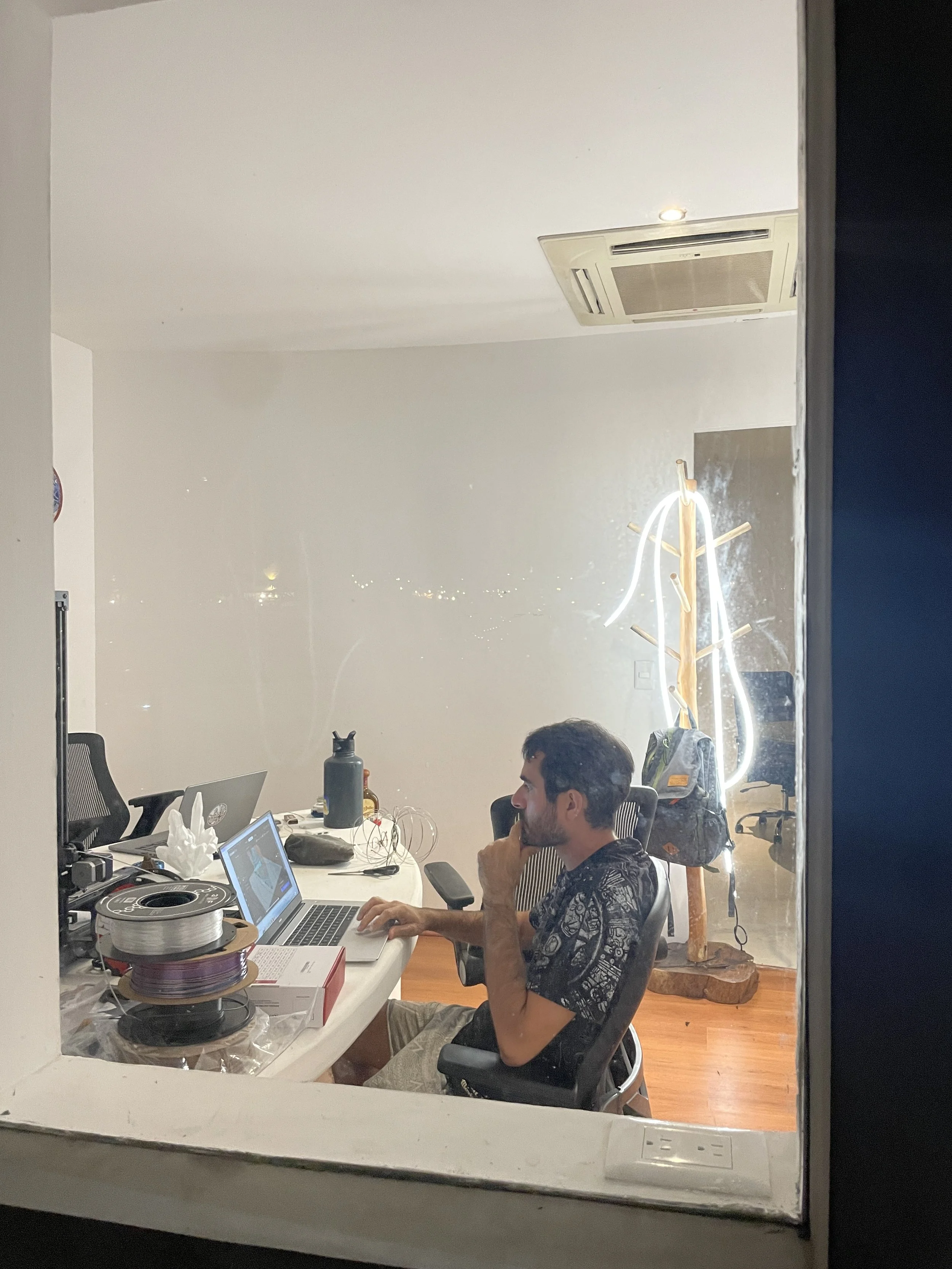 A man sitting at a desk working on a laptop, seen through a window with a reflection of a neon sign outside.