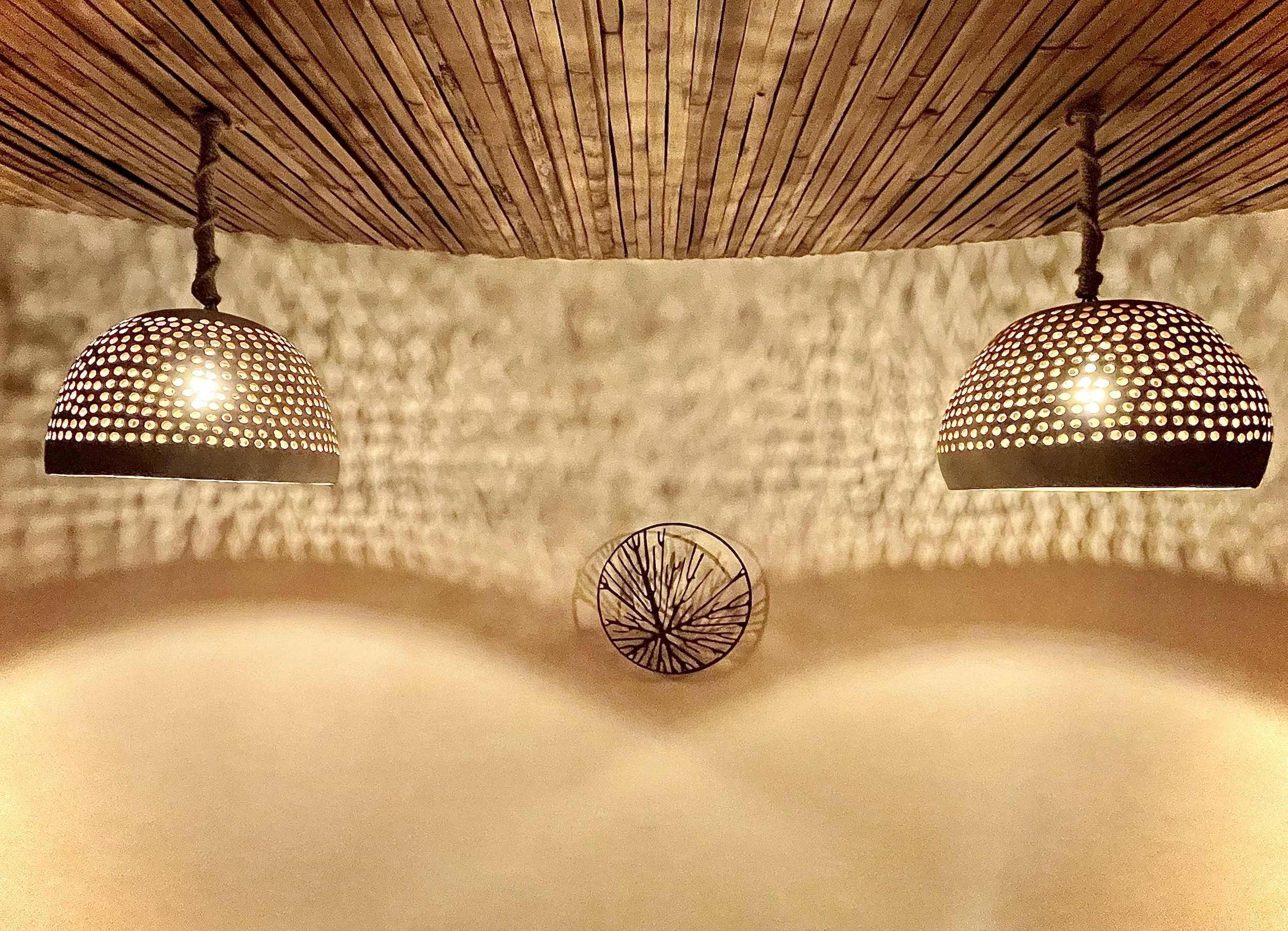 Two pendant lights with perforated shades hanging from a wooden ceiling, casting shadows on a textured wall. A circular decorative piece with a tree branch design is mounted on the wall between the lights. Hypha Inc Marketing Agency Creative Studio