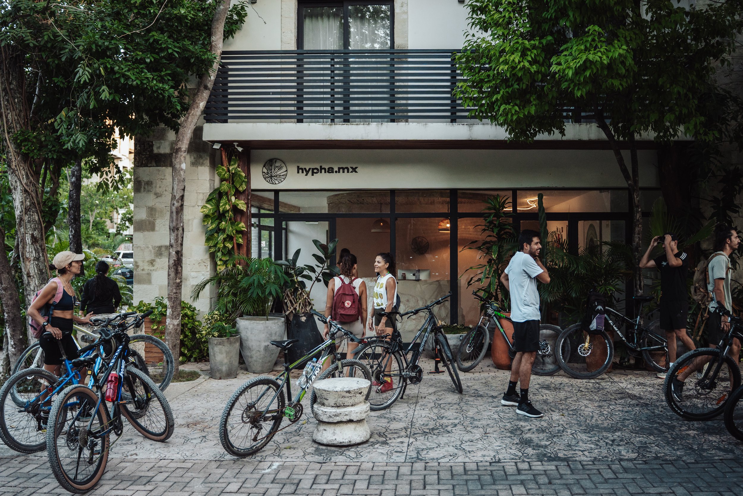 People gathered with bicycles outside a building with a sign reading 'hypha.mx', surrounded by trees and plants.