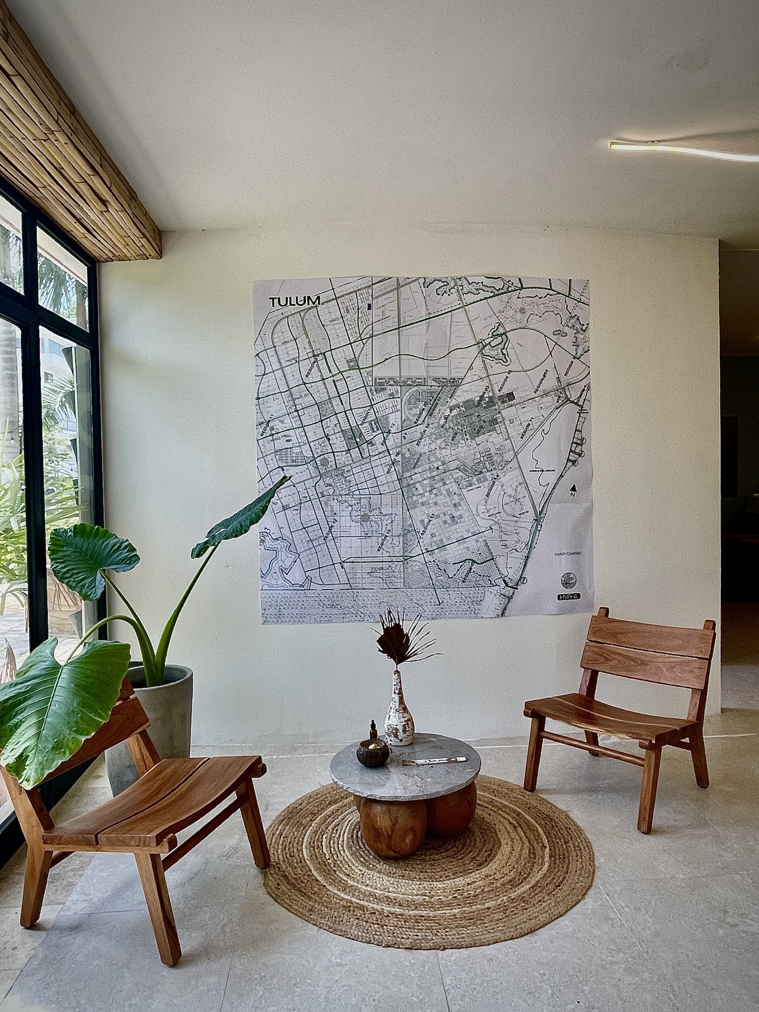 An indoor seating area featuring two wooden chairs, a circular rug, and a small round table with vases, near a large window with greenery outside and a wall map of Tulum. Hypha Inc Marketing Agency and Creative Studio Lab, Tulum.