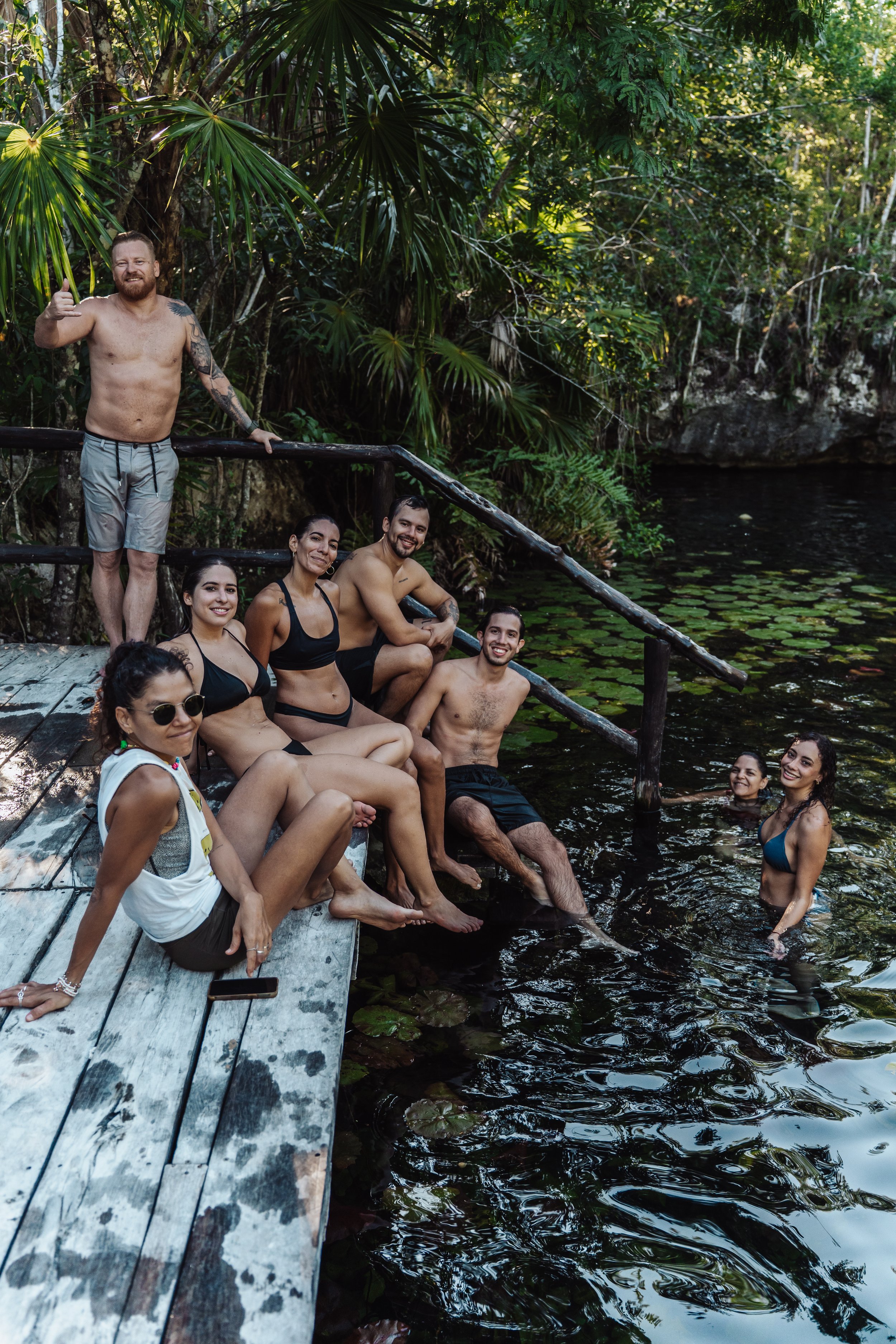 Group of nine people enjoying time by a natural pond in a lush, tropical forest. Some are sitting on a wooden dock, others are in the water. They are smiling and relaxed.