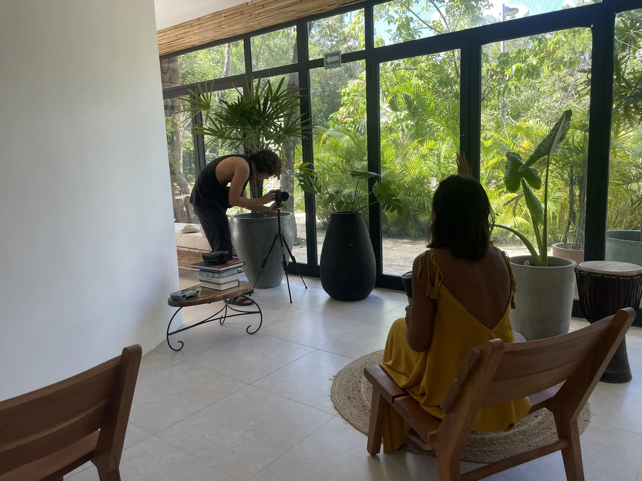 A woman in a yellow dress sits on a wooden chair in a bright room with large glass windows, while a man adjusts a camera on a tripod nearby, with lush green plants outside.