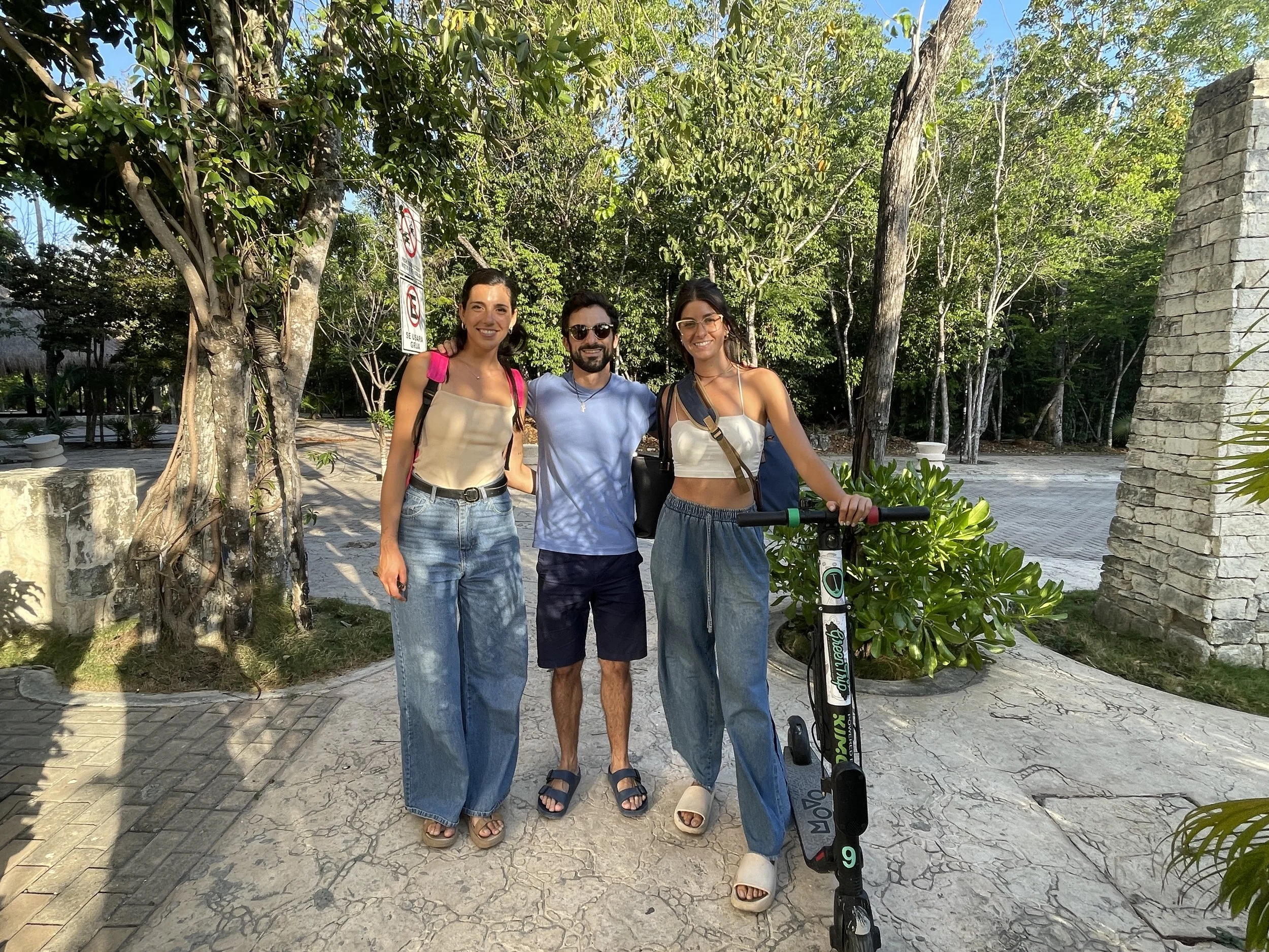 Hypha Team standing outdoors on a sunny day, smiling at the camera. Two women and one man, with trees and a stone wall in the background. The woman on the right is holding an electric scooter. Hypha Inc. Digital Marketing & Advertising Agency
