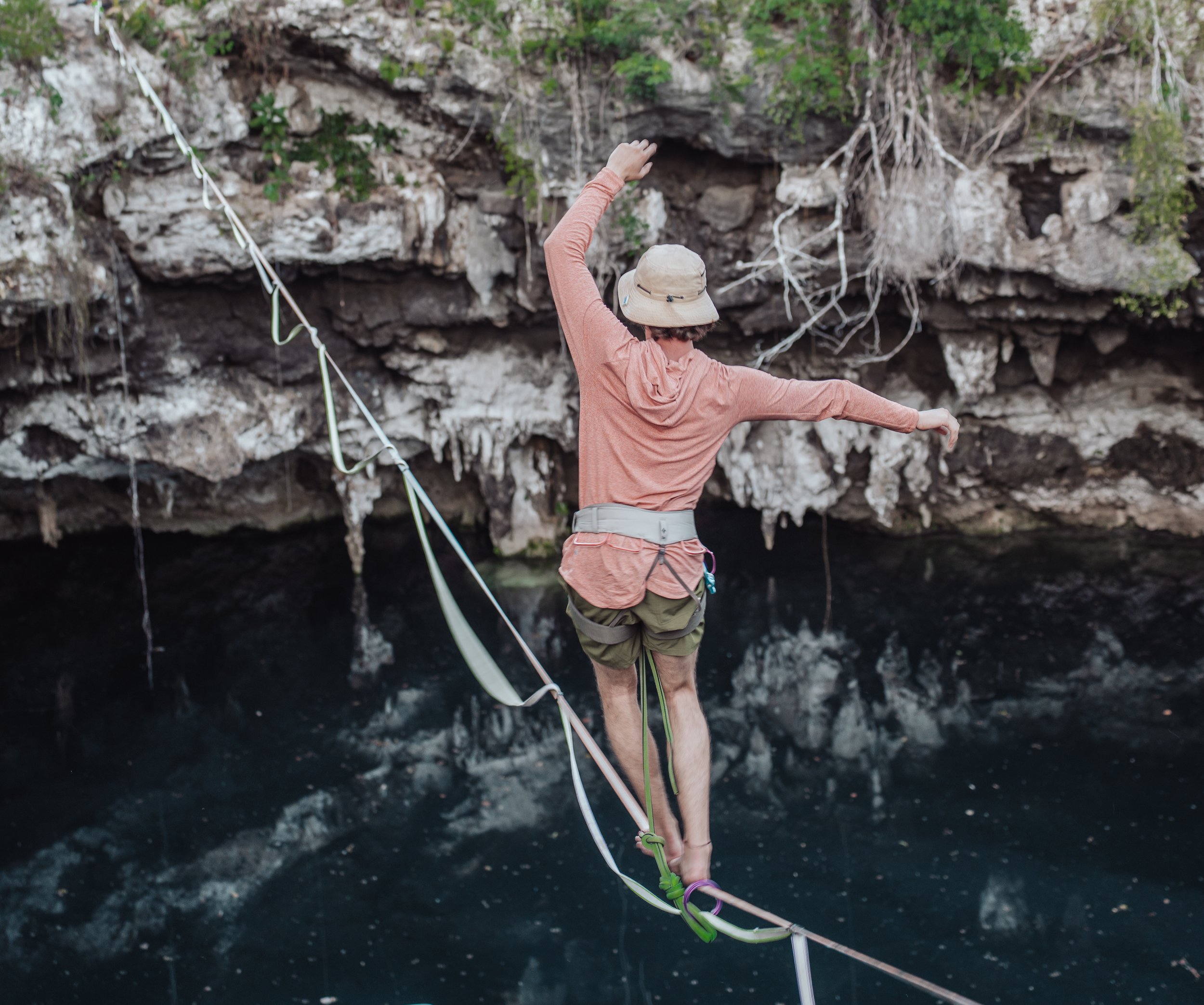 Person wearing a hat and outdoor clothing balancing on a slackline over water near a rocky cliff in Cenote in Riviera Maya