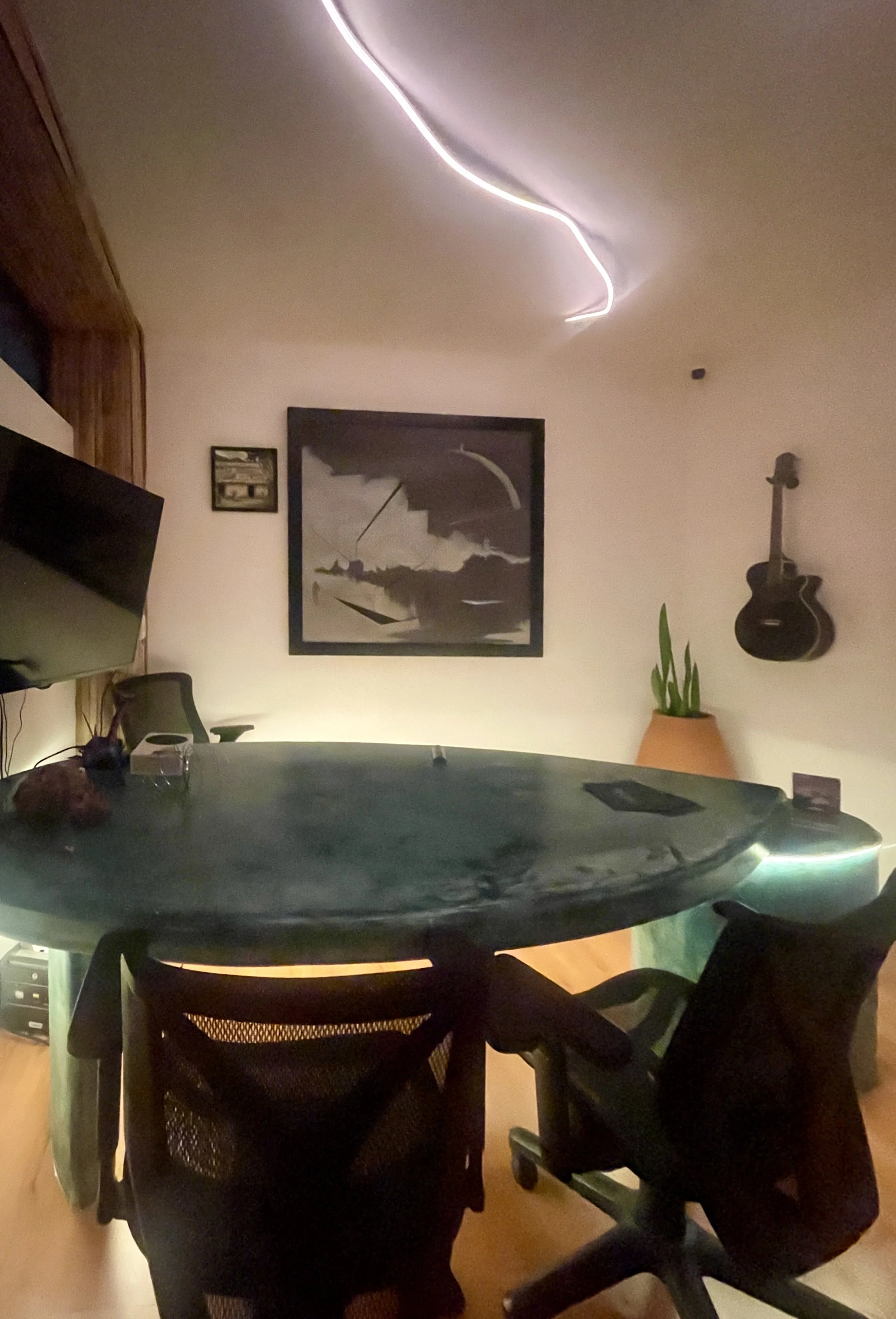Office with a round table, two black office chairs, a wall-mounted TV, a framed black-and-white drawing, a guitar on the wall, a potted plant, and ceiling lights. Hypha Inc. Digital Marketing & Advertising Agency, Tulum