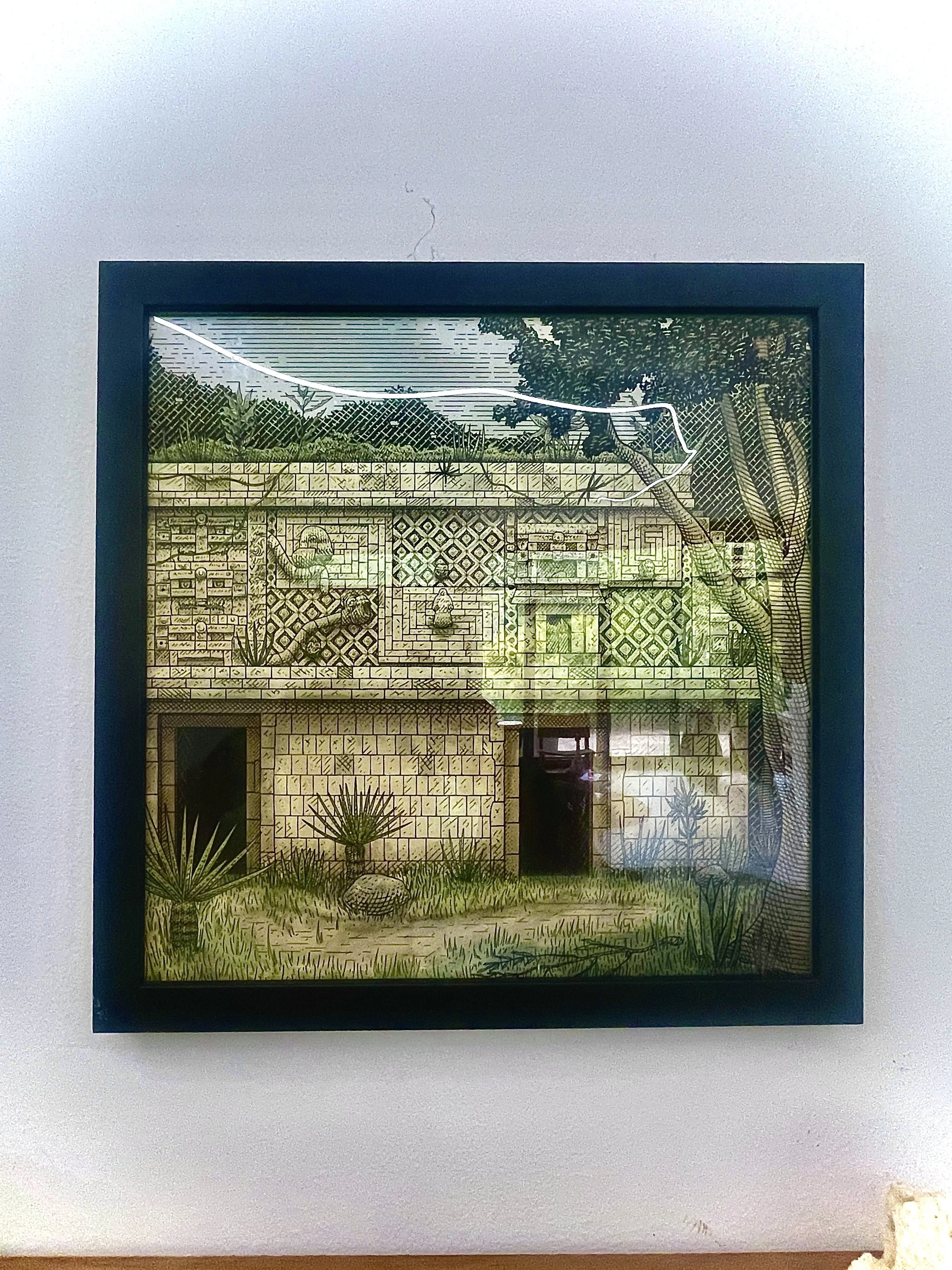 Framed artwork of an ancient stone building with geometric patterns, surrounded by desert plants and trees, displayed on a white wall.