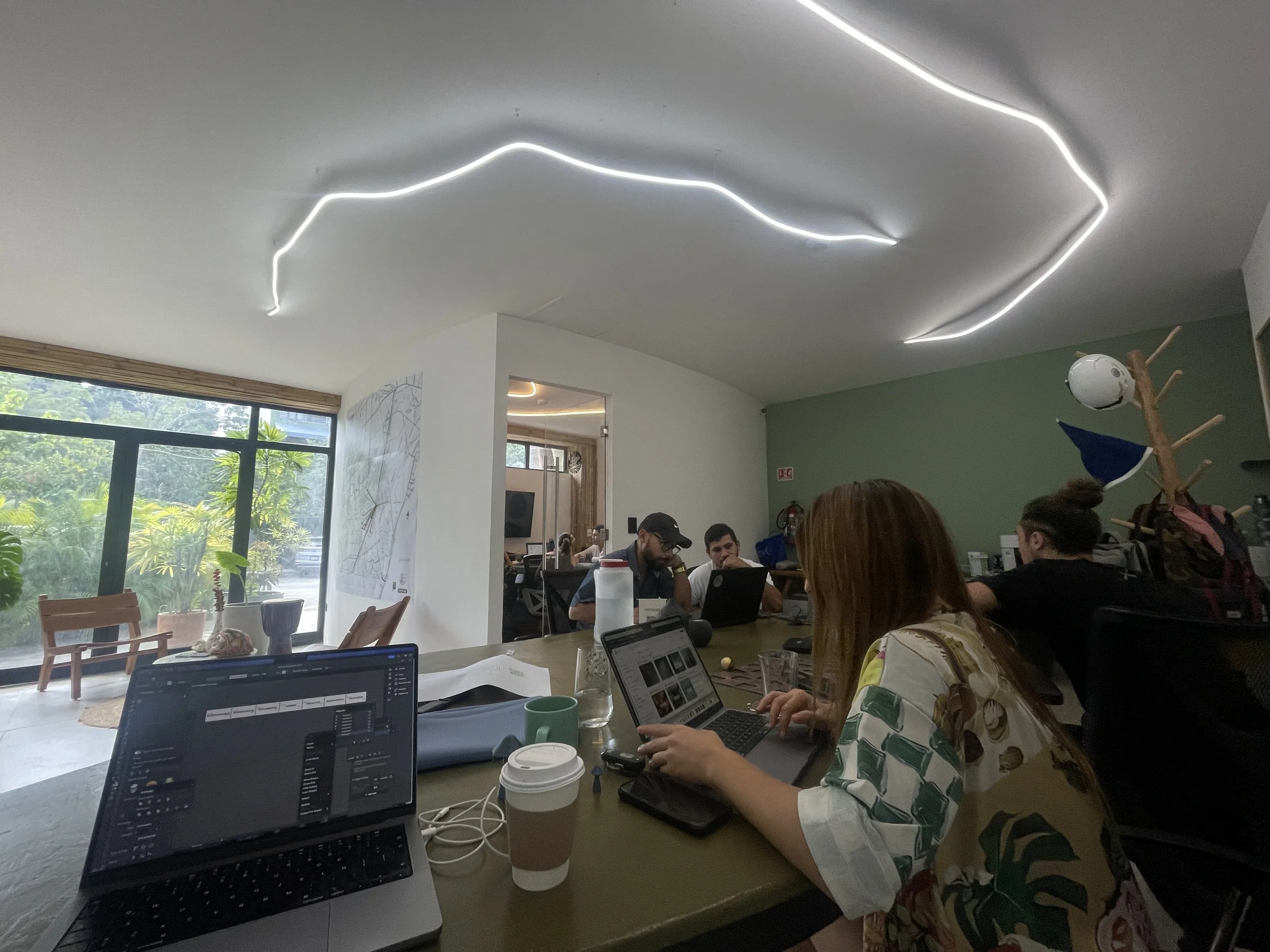 People working and collaborating around a conference table in a modern office space with large windows and greenery outside. Laptops, coffee cups, and office supplies are visible on the table. The ceiling has unique curved LED lighting in Hypha Inc 