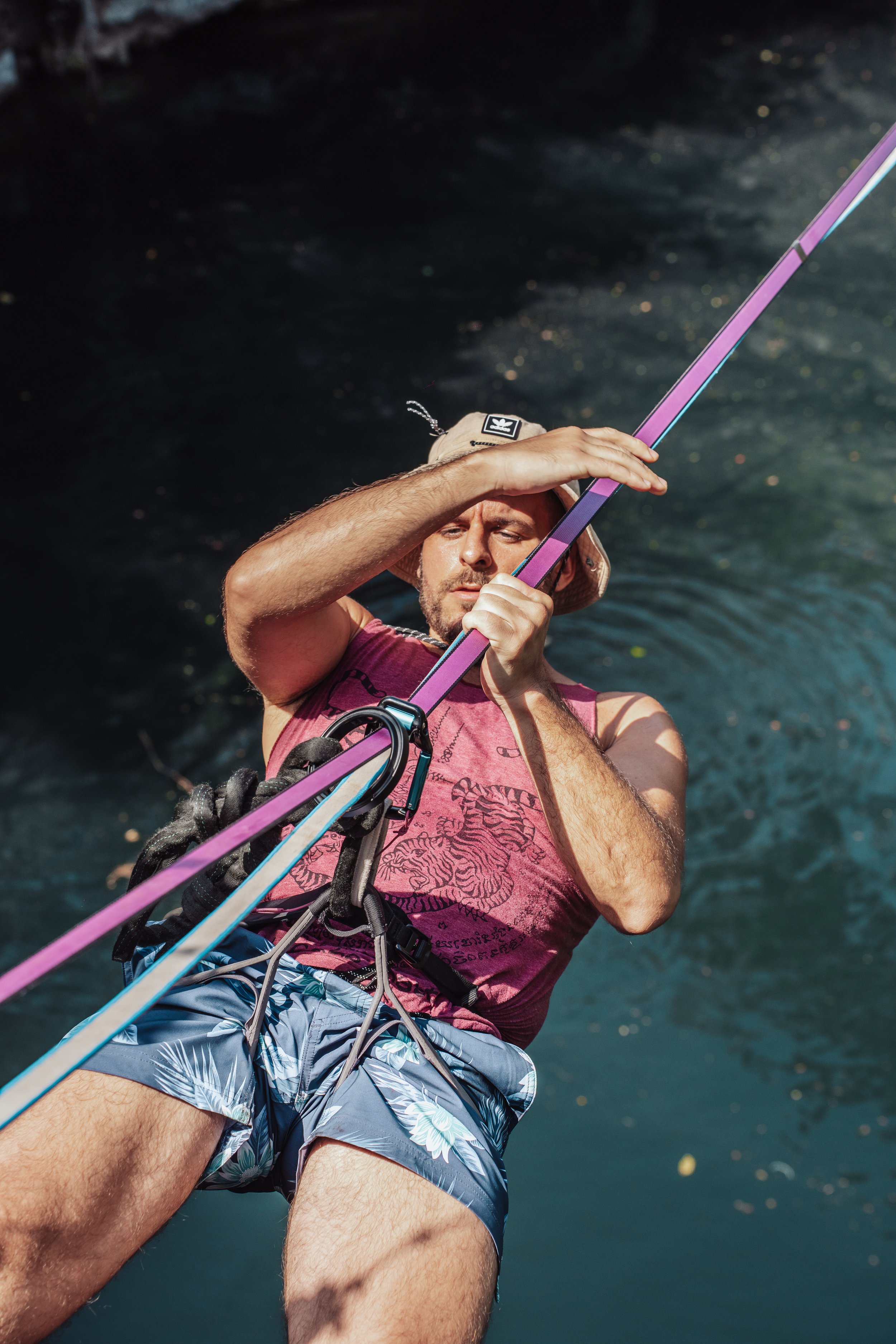 A man wakeboarding outdoors on a sunny day, holding a wakeboard rope with a purple handle, wearing a sleeveless shirt, floral shorts, and a bucket hat, with cenote cave in the background.