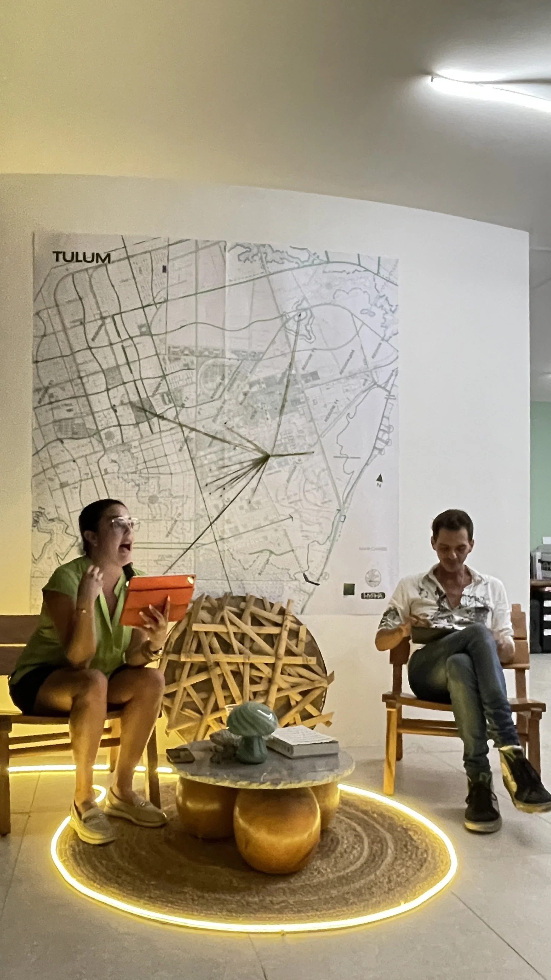 Two people sitting in chairs in front of a large map on the wall. The person on the left is a woman holding an orange tablet, appearing to be speaking or explaining something, while the person on the right is a man looking down at a book or notebook.