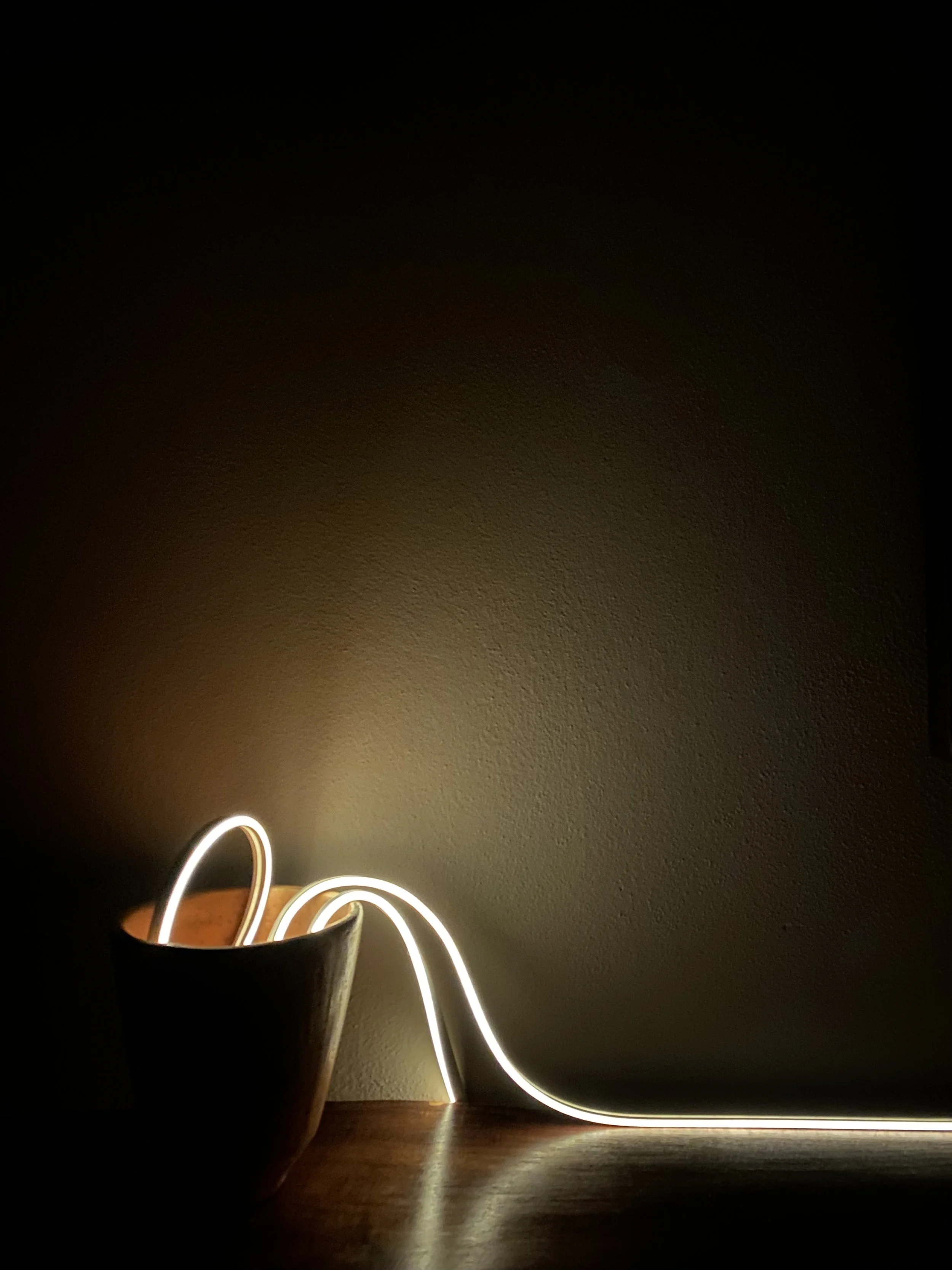 A black bowl with a glowing white LED light strip inside, creating a curvy line that extends out of the bowl and runs along the wall behind it, illuminating the beige wall and wooden surface. Hypha Inc Marketing Agency and Creative Studio Lab, Tulum.