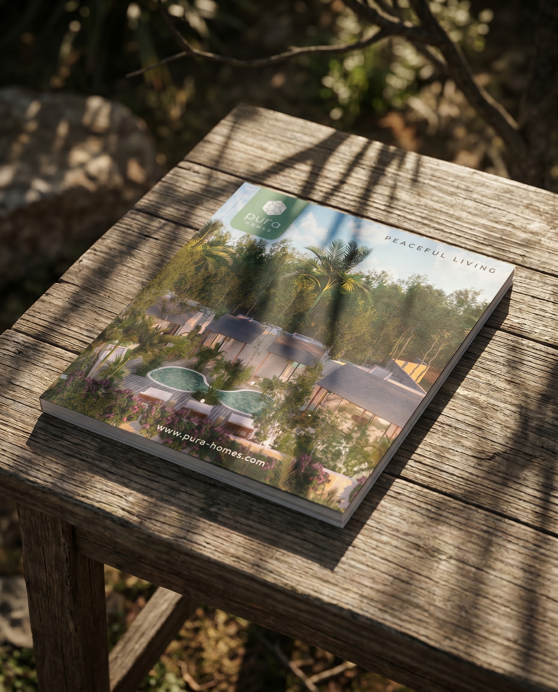Brochure or magazine with an image of a modern house surrounded by trees and palm trees, placed on a weathered wooden table outdoors. The brochure features the logo 'pura homes' and the text 'peaceful living'. Hypha Inc. Branding and Design Agency