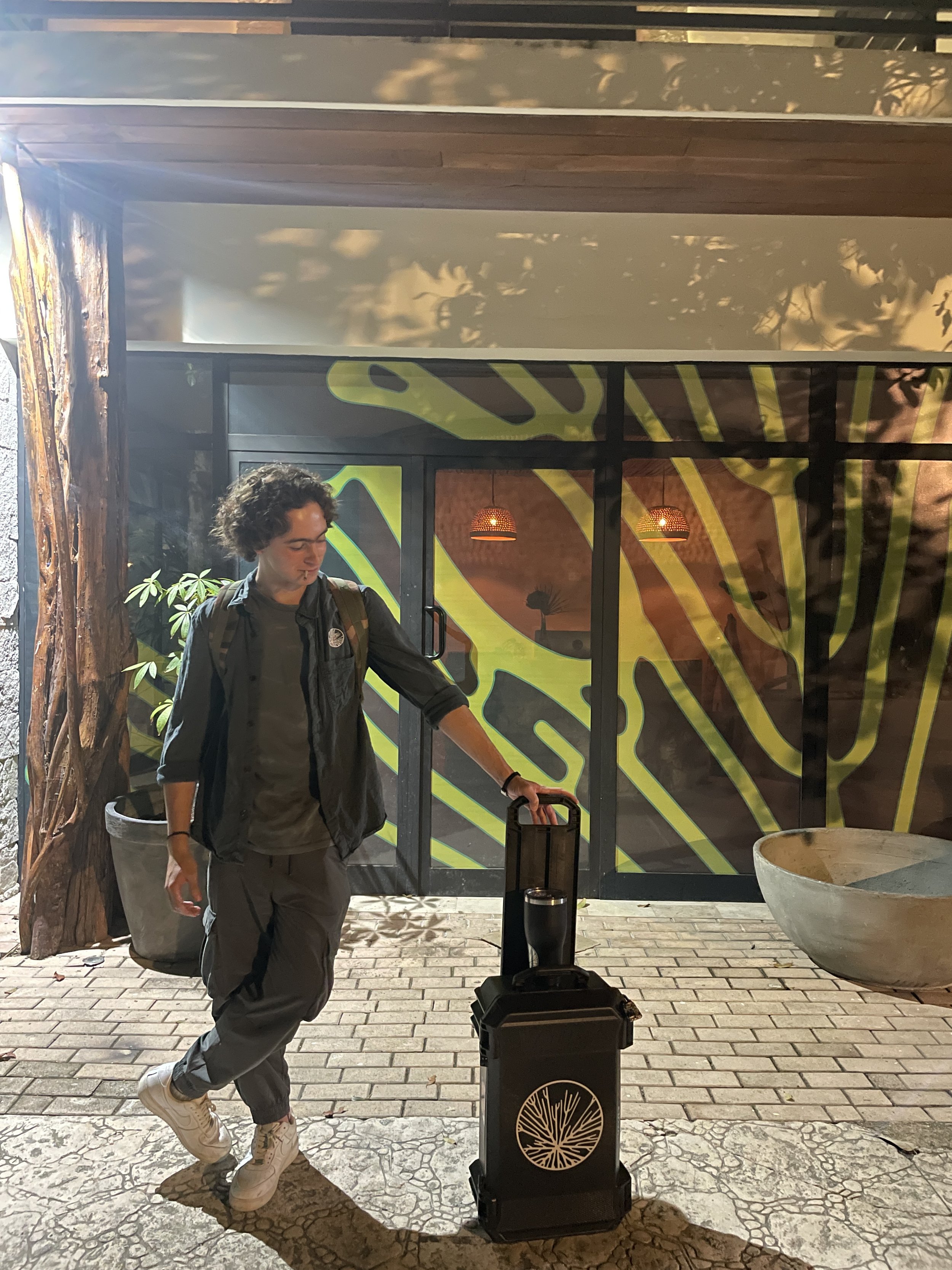 Pato, dressed in casual Hypha tshirt, standing outside at night, holding a rolling suitcase, with a colorful, abstract mural on the glass door behind him, and a large concrete planter nearby.