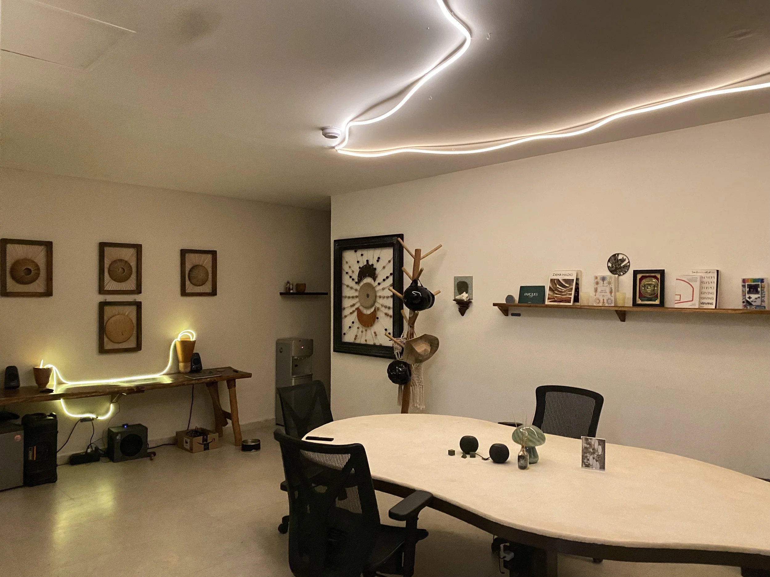 This is an indoor room with a white wall, ceiling lighting, a table with chairs, decorative art pieces, a wooden coat rack, and shelves with books and decorative items. Hypha Inc Marketing Agency and Creative Studio Lab, Tulum.