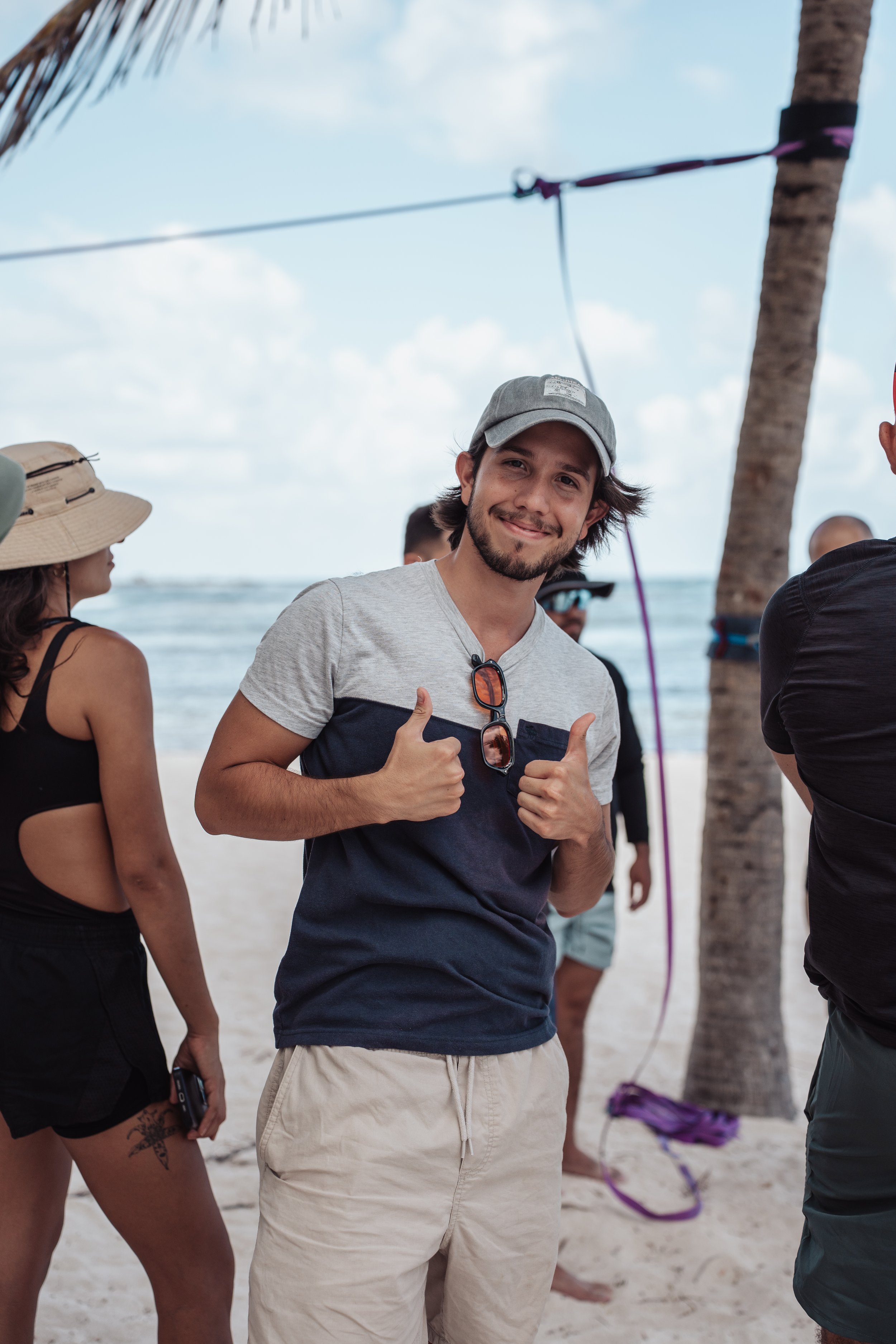 A smiling young man with a beard giving a thumbs up while standing on a beach with palm trees, other people, and the ocean in the background.