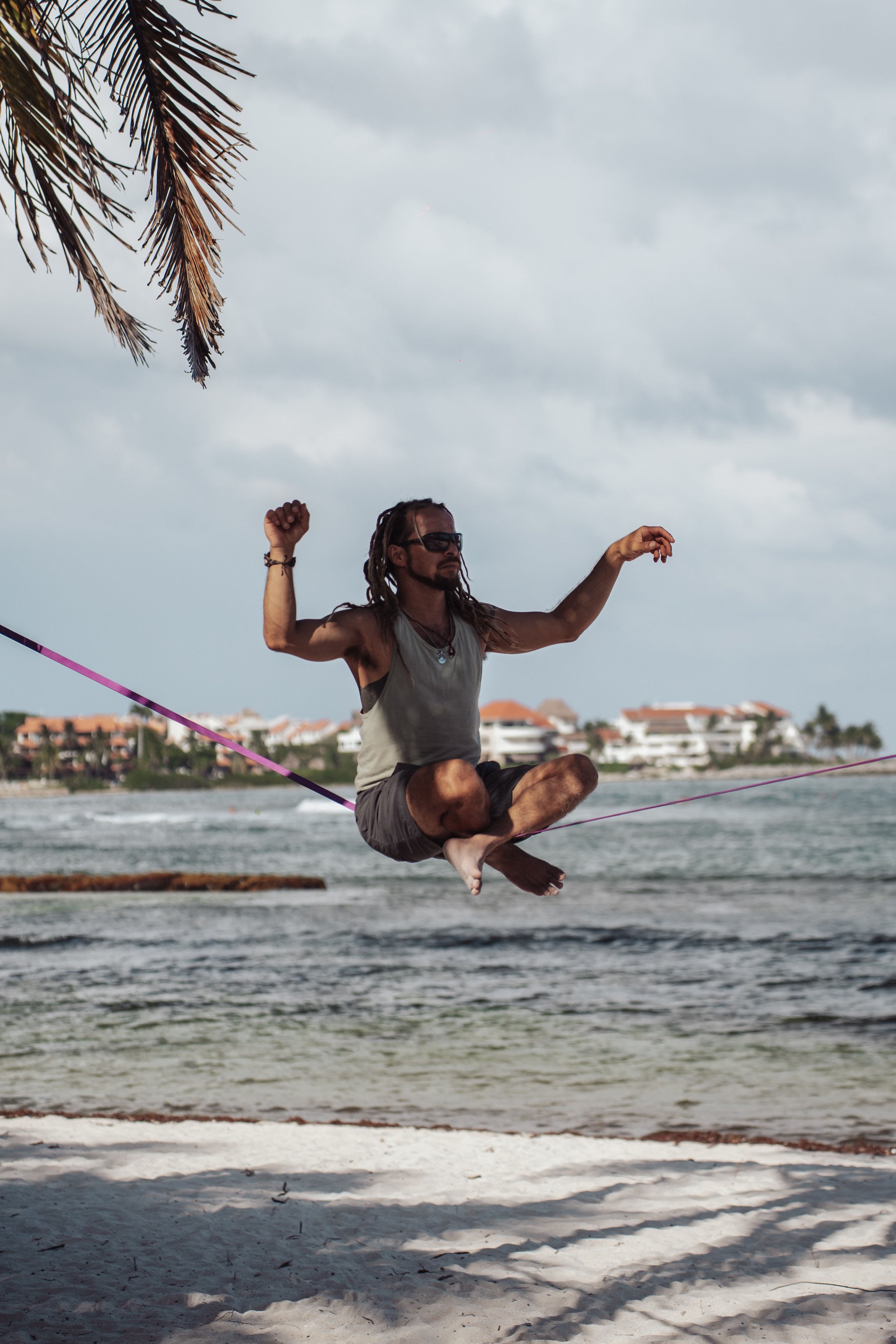 A man with long hair and sunglasses is sitting cross-legged on a slackline string, balancing above the sandy beach near the ocean, with buildings and palm trees in the background under cloudy skies.