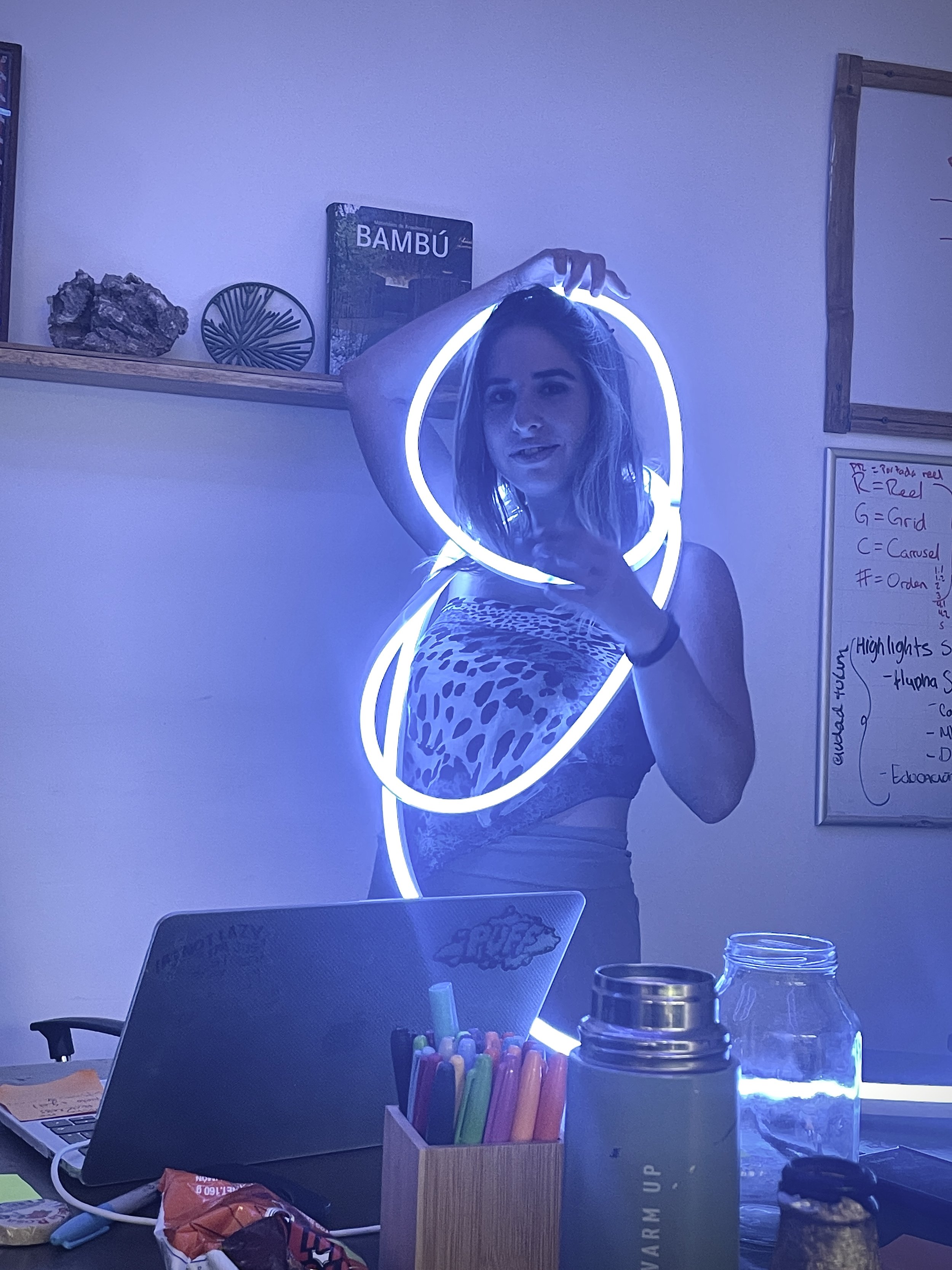 APOD standing in front of a whiteboard, wearing a unique outfit made of illuminated neon light rings. The photo is taken indoors with office supplies on a desk in the foreground. Hypha Inc Marketing Agency and Creative Studio Lab, Tulum.
