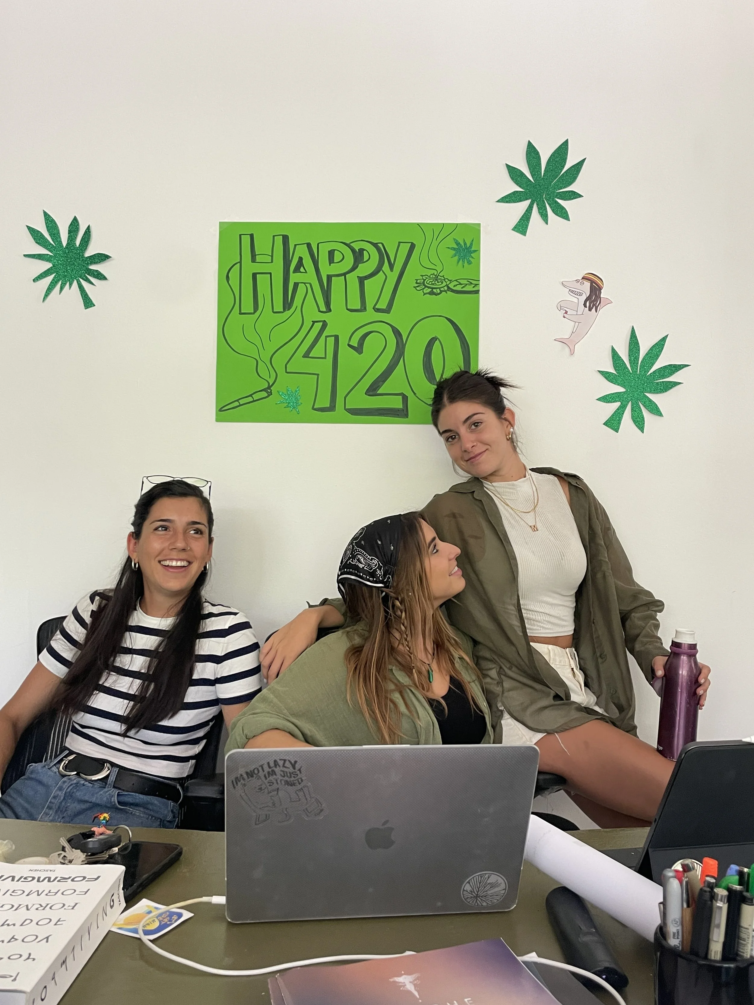 Three women in an office celebrating a 420-themed birthday, with a green "Happy 420" poster and marijuana leaf decorations on the wall. Hypha Inc Marketing Agency and Creative Studio Lab, Tulum.