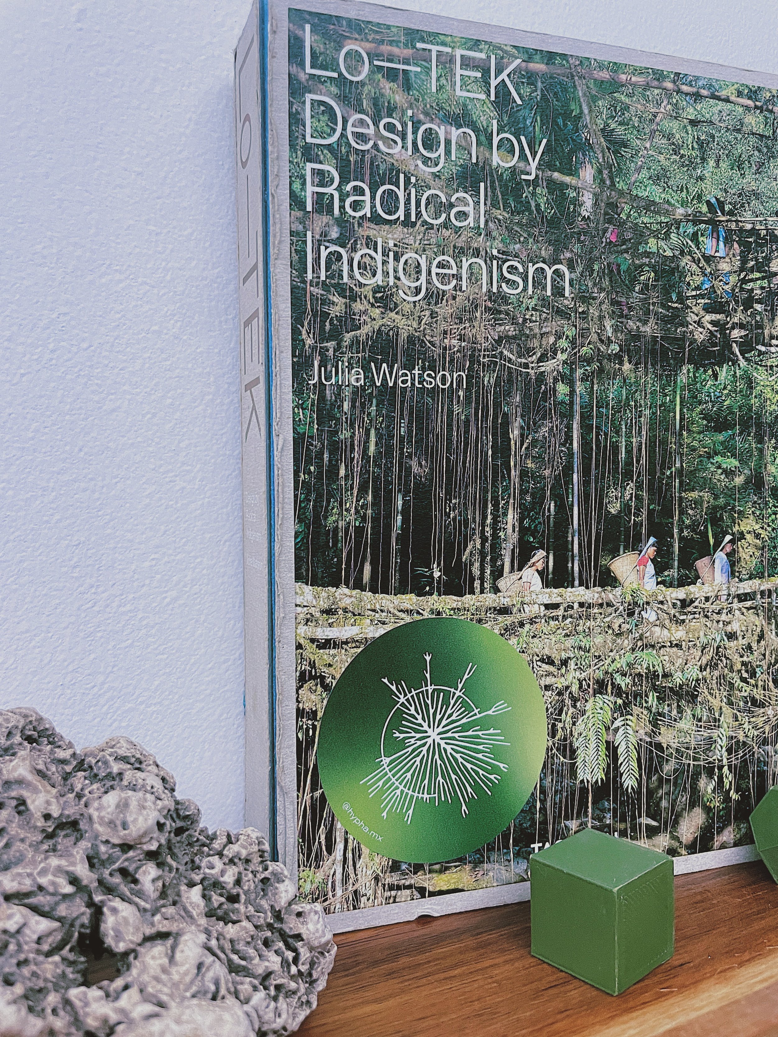 Book titled 'Lo+Tek: Design by Radical Indigenousism' by Julia Watson, featuring a forest scene with indigenous people carrying baskets on a bridge on the cover. Hypha Inc Marketing Agency and Creative Studio Lab, Tulum.
