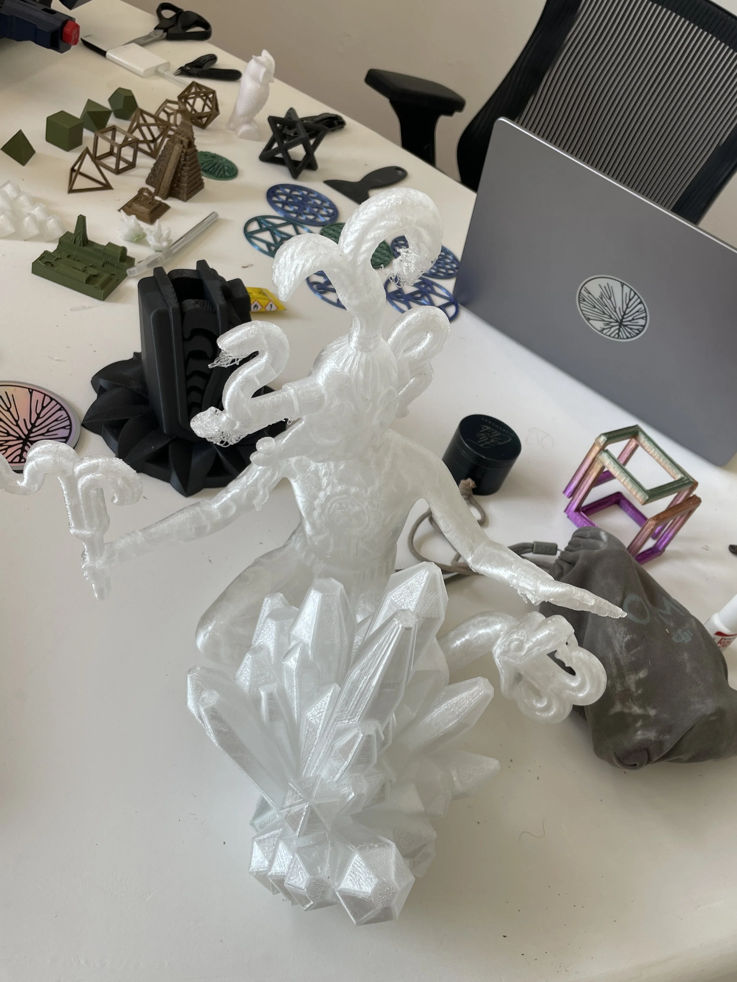 Translucent 3D-printed sculpture of a mythical bird standing on crystal-like base, placed on a white table with various small objects and 3D prints scattered around.